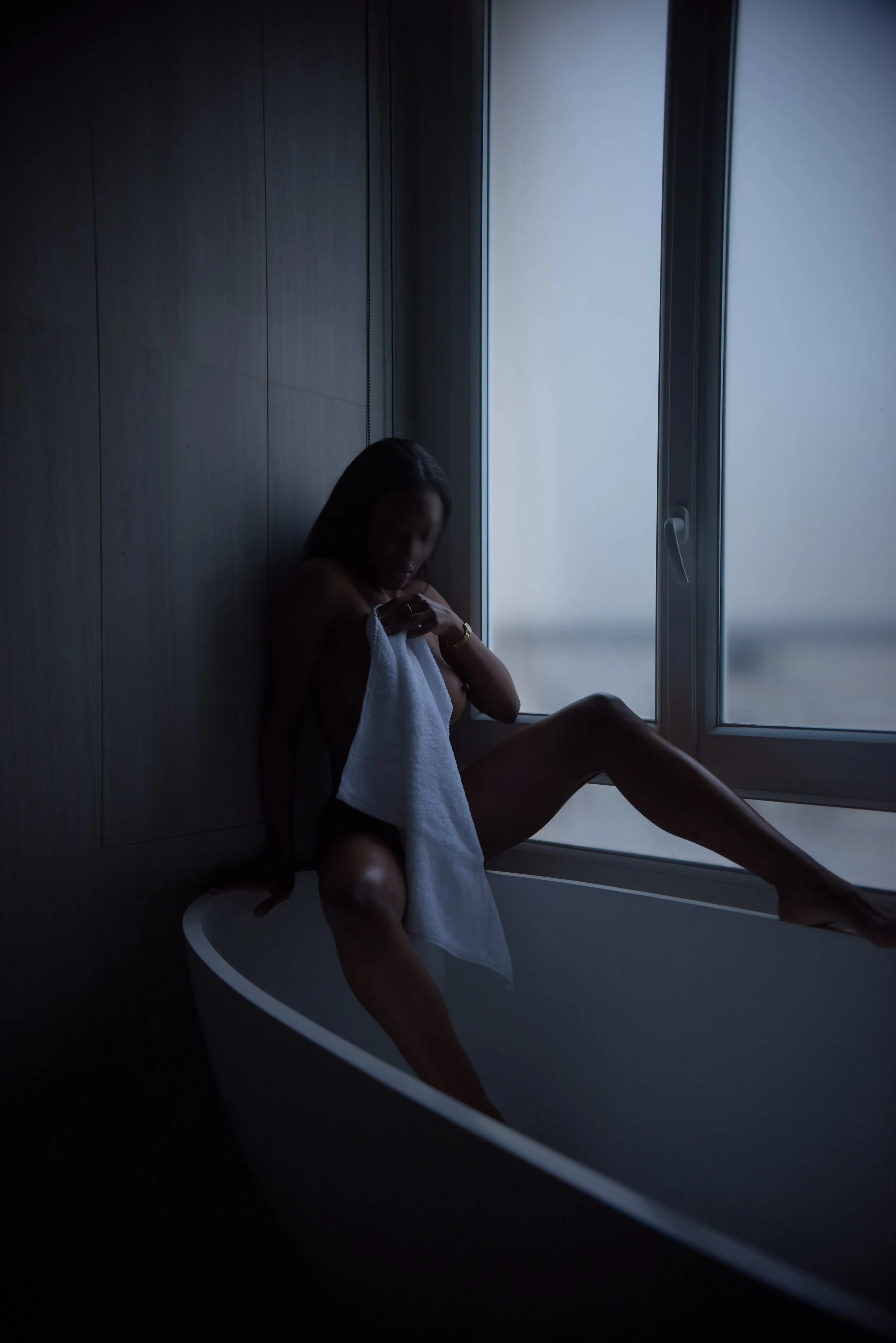 Femme nue assise dans une baignoire près d'une fenêtre, tenant une serviette blanche, dans une pièce sombre avec un éclairage naturel faible.