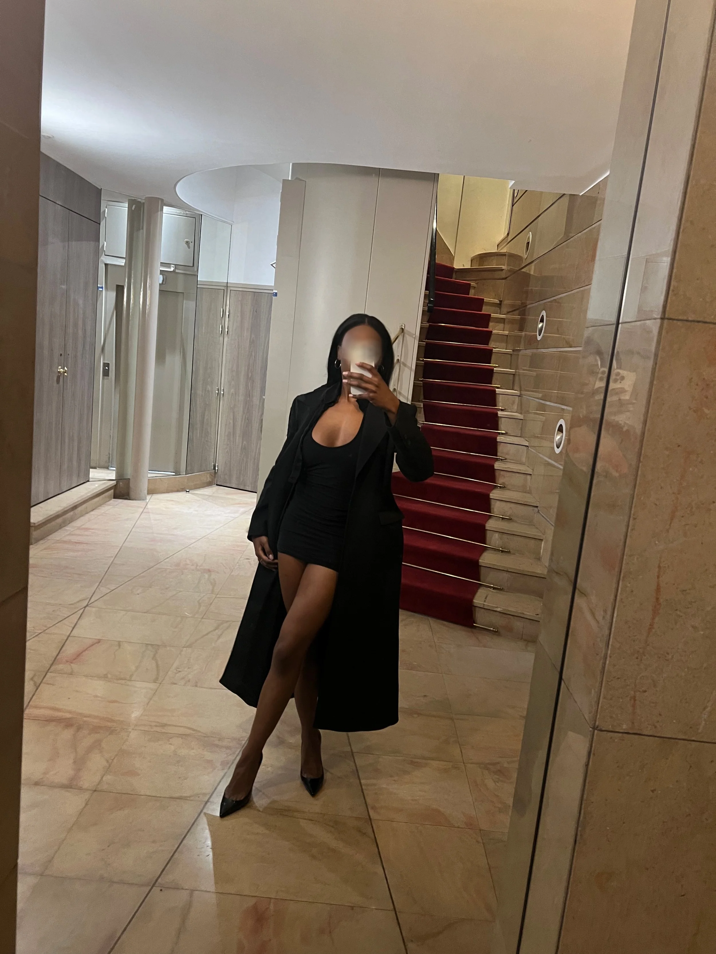 Une femme habillée en noir, portant un trench et une robe courte, se prenant en selfie devant un miroir près d'un escalier dans un bâtiment.