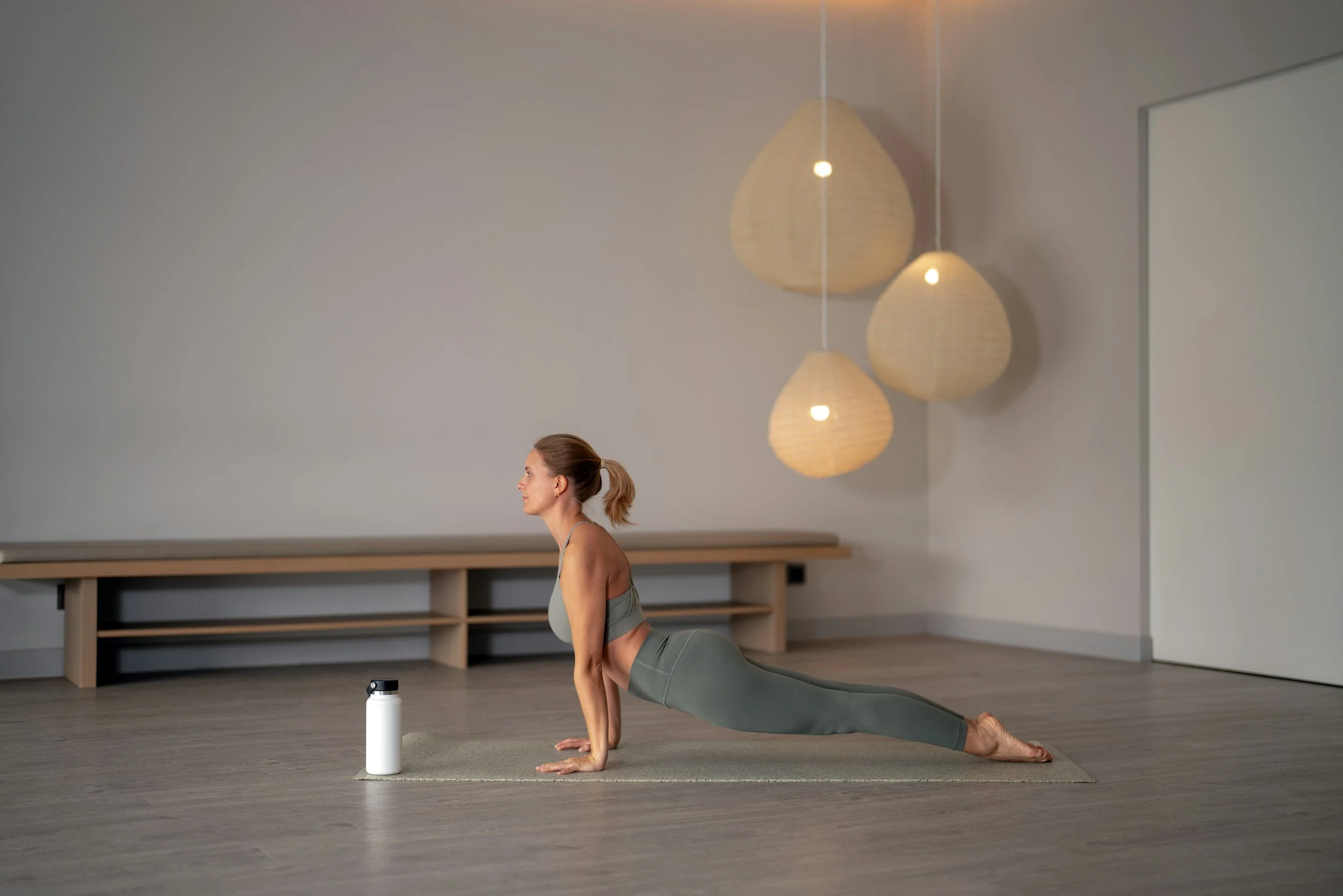 Woman practicing yoga in a minimalist studio with hanging paper lanterns, performing cobra pose on a yoga mat with a water bottle nearby.