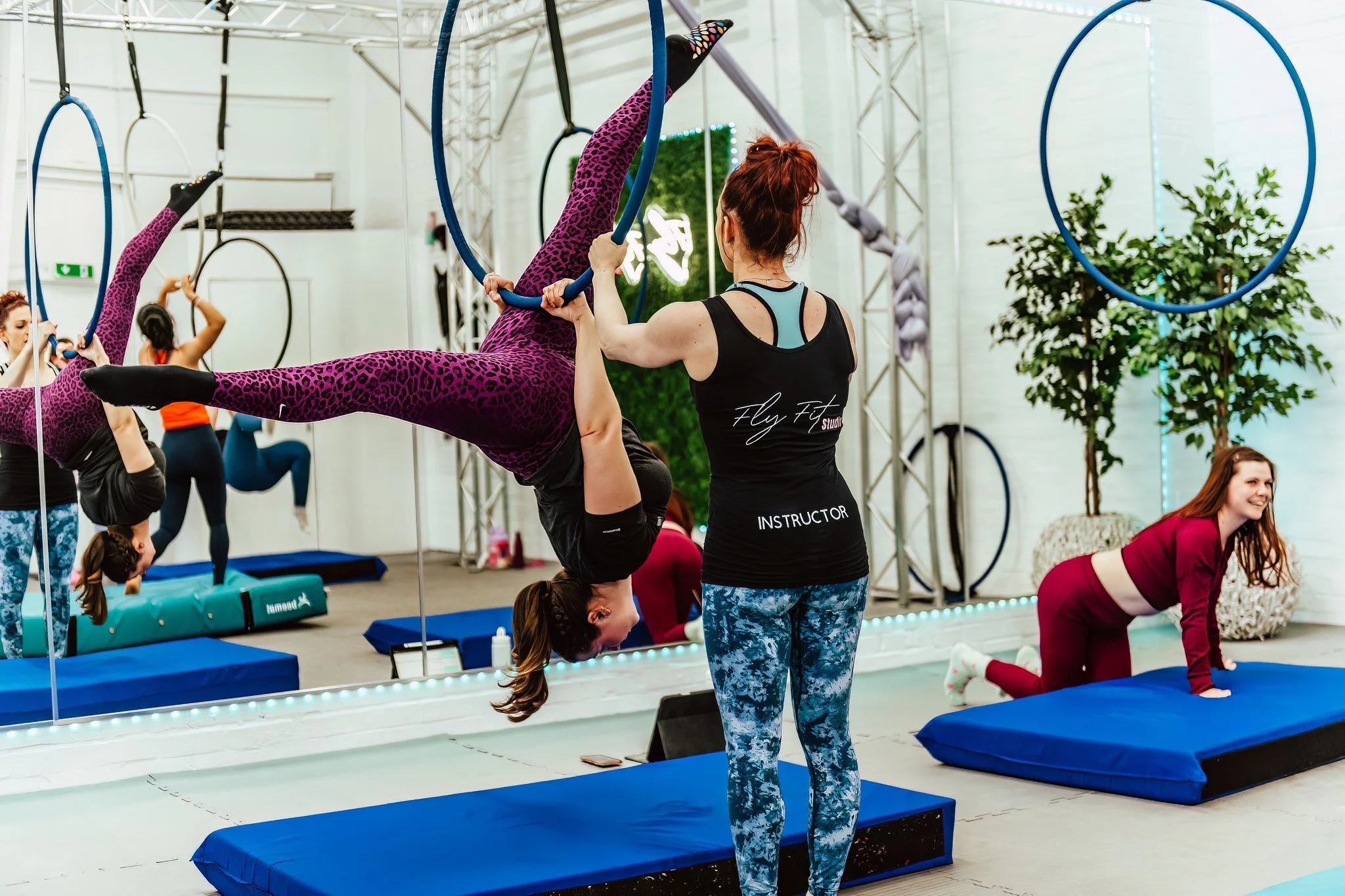 Aerial fitness class with participants using hoops and mats, instructor guiding a woman in purple leopard print leggings in an upside-down pose, studio with mirrors and plants.