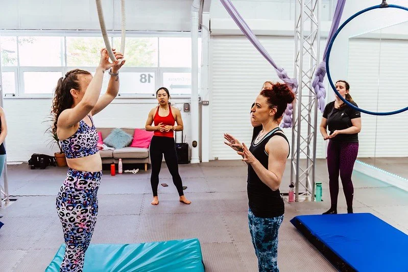 A fitness instructor giving instructions to women in a gym, with one woman hanging from aerial silks and others standing nearby.