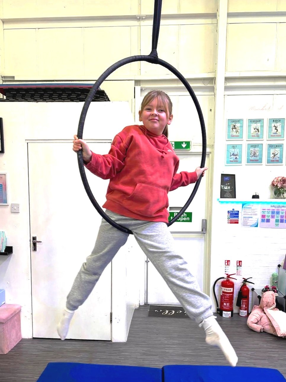 A young girl in a red hoodie and gray pants jumping on a blue mat while holding a black hanging hoop at an indoor recreational center.