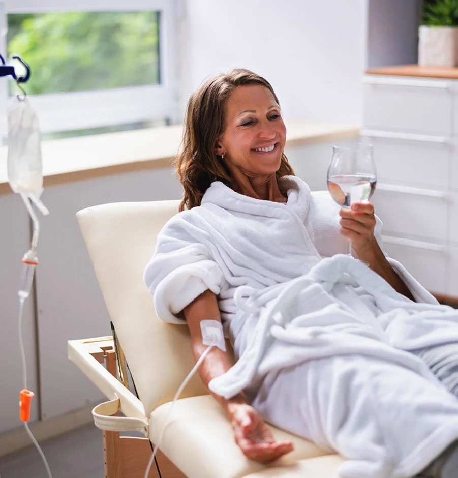 Smiling woman in a bathrobe relaxing with a glass of water during an in-home mobile IV therapy session