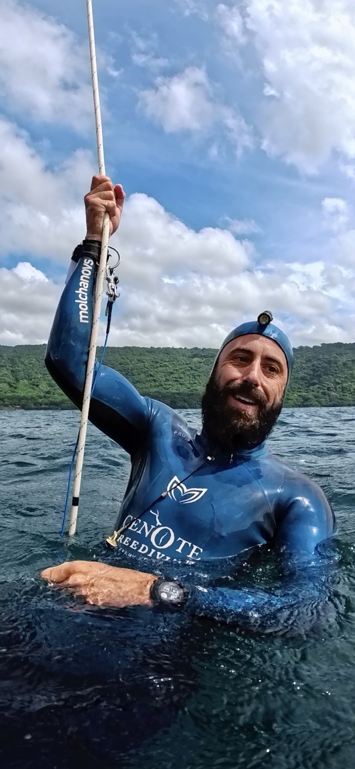 A man in a blue wetsuit with a headlamp, holding an underwater pole, smiling while standing in a body of water with a green forested shoreline in the background.
