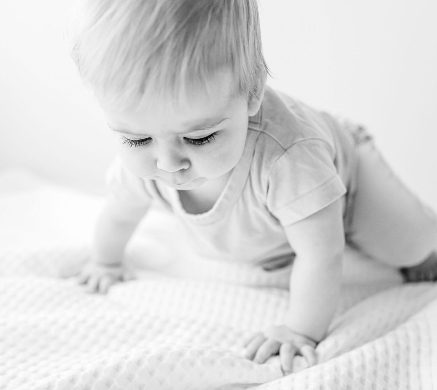 A young child, crawling on a textured blanket, looking downward with focused expression.