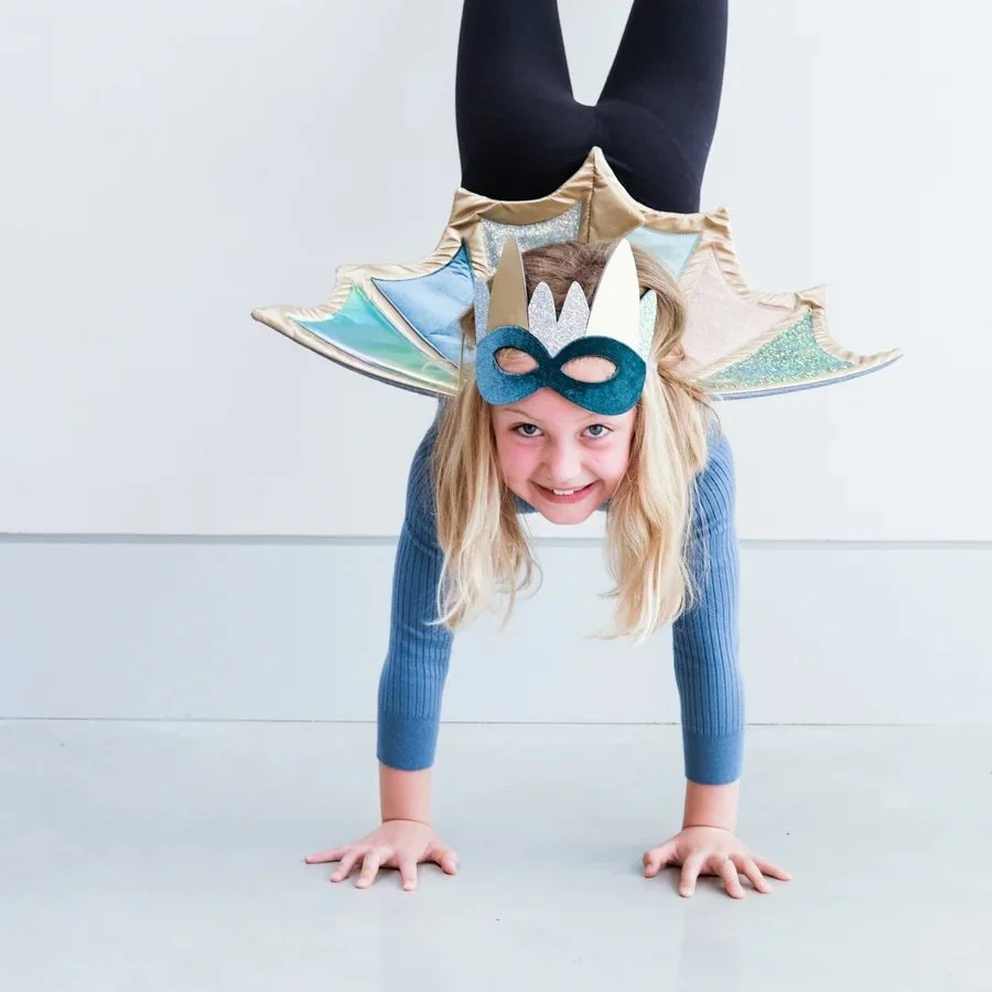 A young girl in a costume doing a handstand with a colorful fairy wing and mask, with her legs extended upward.