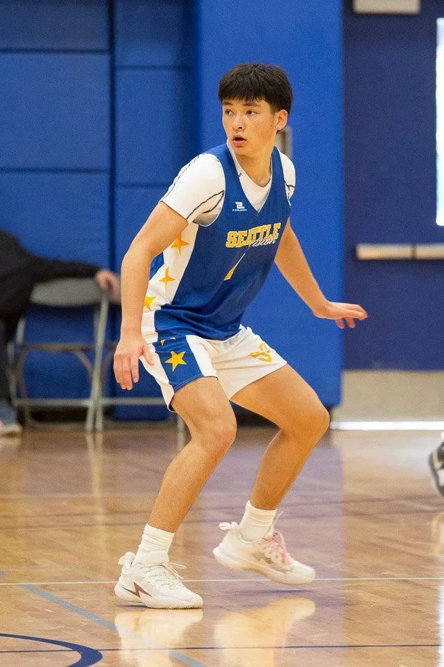 Young male basketball player in a blue and white uniform with yellow stars, playing on a wooden court.
