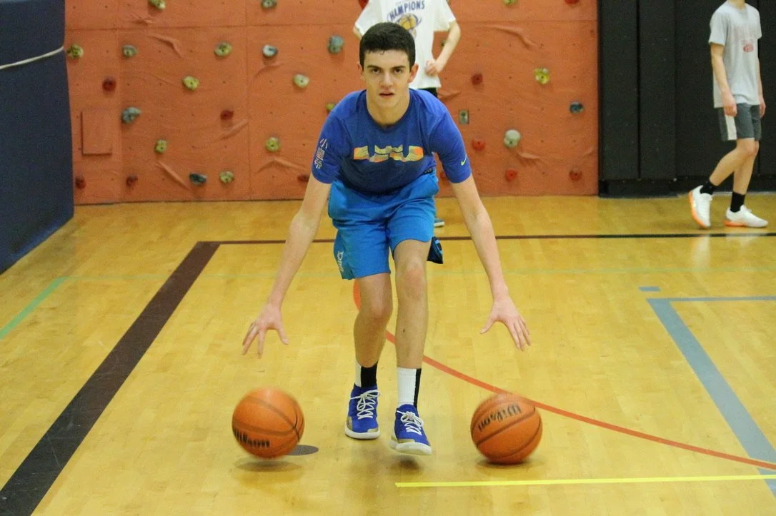 A young man in a blue athletic outfit practicing dribbling two basketballs in a gym with a climbing wall in the background.