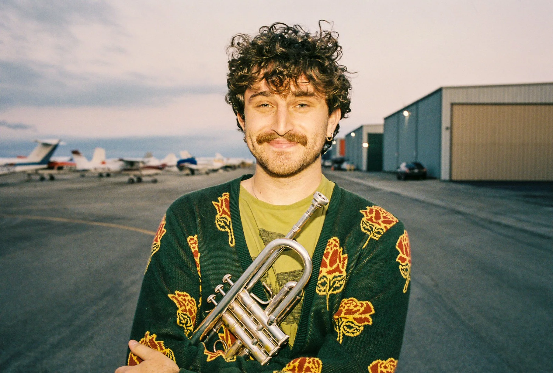 A man with curly hair, a mustache, and a beard, smiling and holding a trumpet, standing on an airport tarmac with airplanes and hangars in the background.