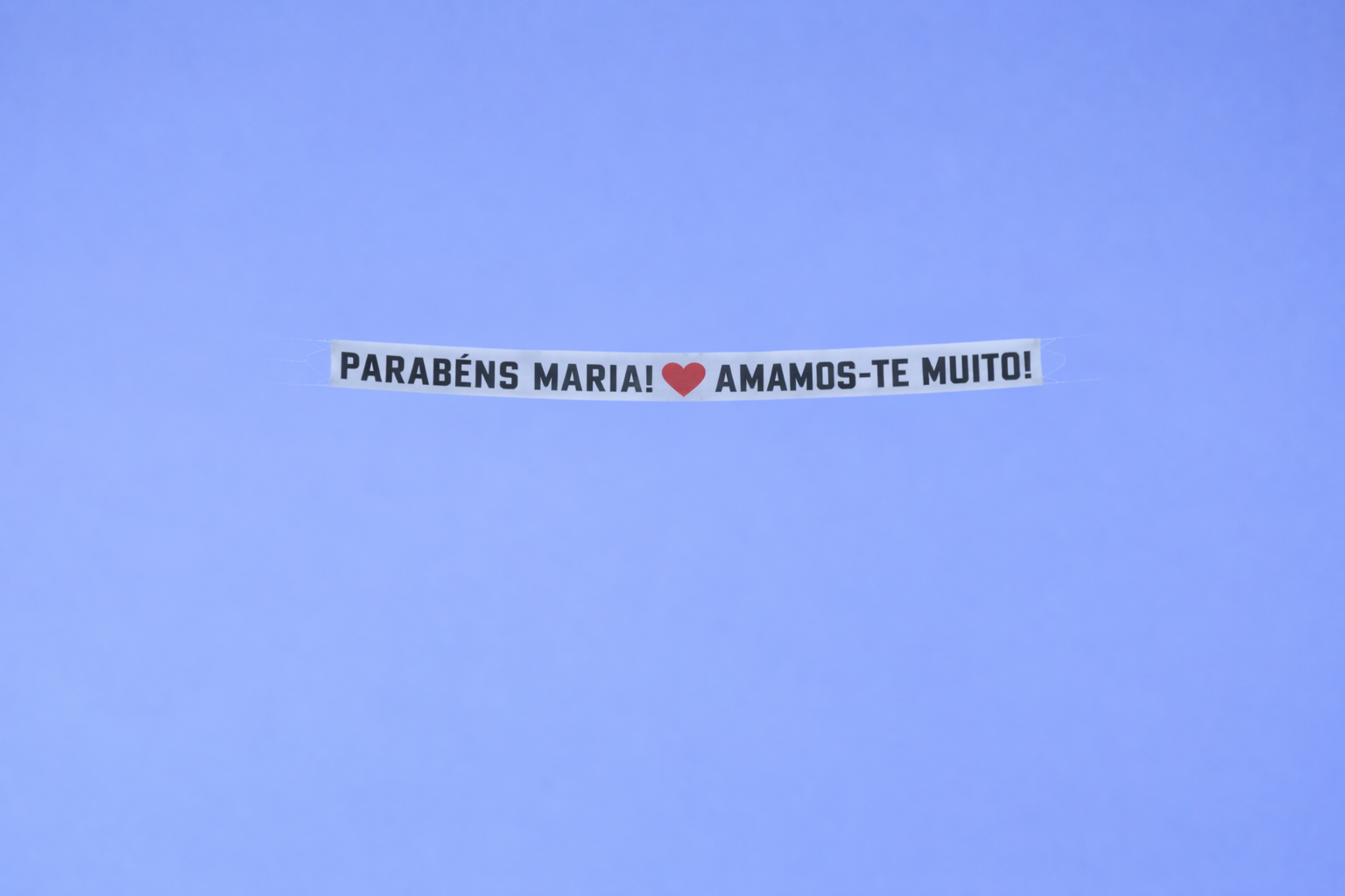 A banner in the sky with the message "Parabéns Maria! Amamos-te muito!" and a red heart symbol between the words.
