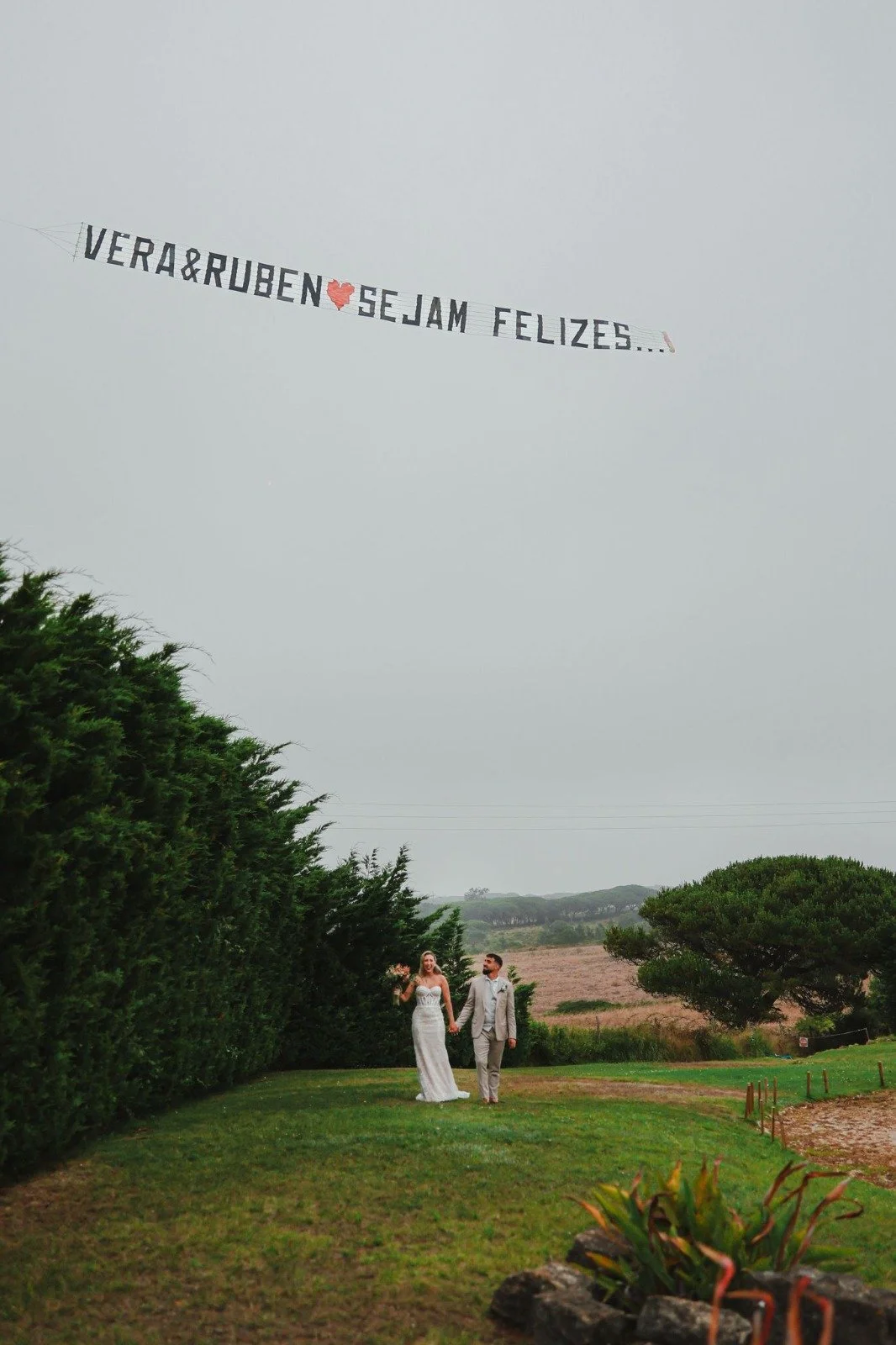 A bride and groom holding hands and walking on a grassy lawn outdoors with a large sign in the sky reading 'Vera&Ruben ❤️ Sejam Felizes...'.