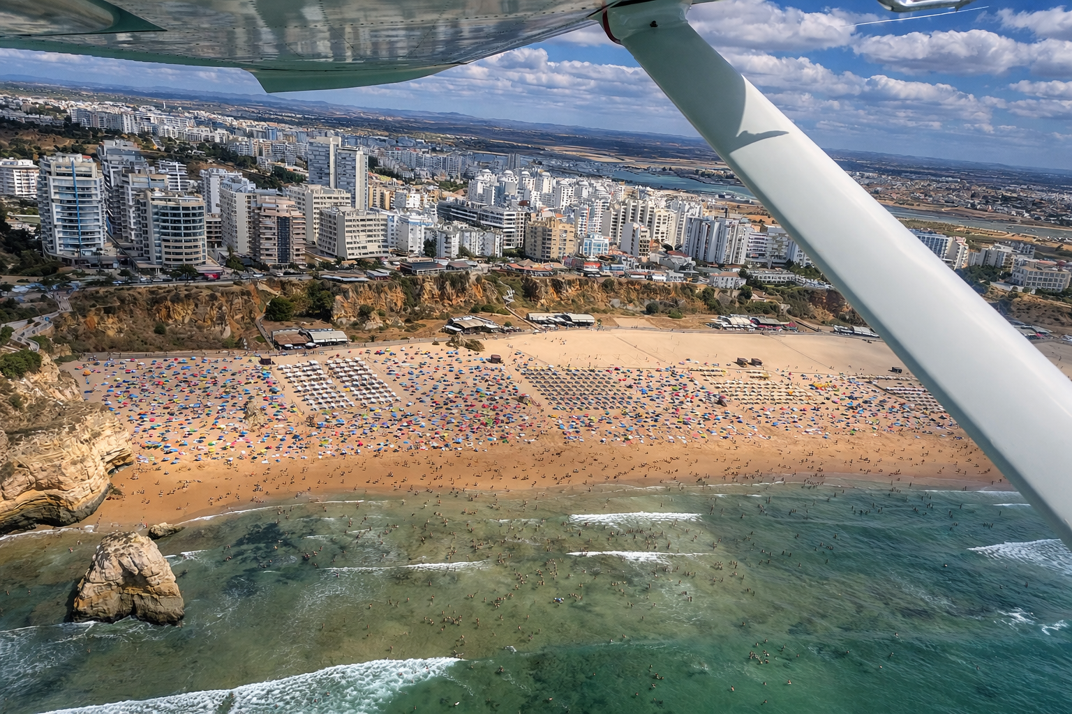 Aerial view of a crowded beach with umbrellas and umbrellas along the sandy shoreline, with many people swimming and surfing in the water. In the background, there are high-rise buildings and a cityscape under a partly cloudy sky, and part of an airplane wing is visible in the foreground.
