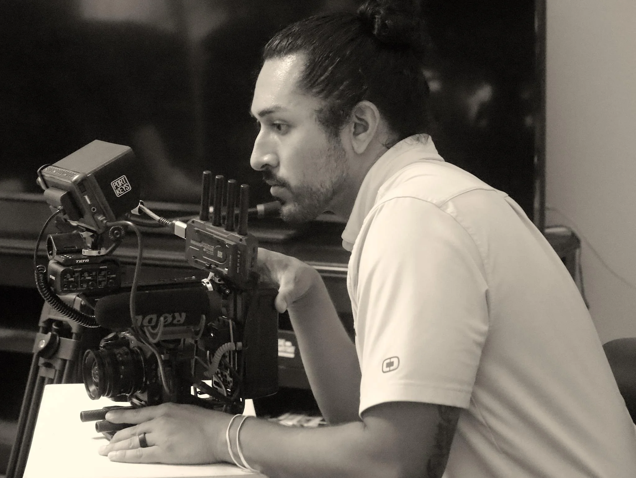 A man with long hair tied back, wearing a short-sleeved shirt, is operating advanced film or video production equipment on a table, focused intently on the work.