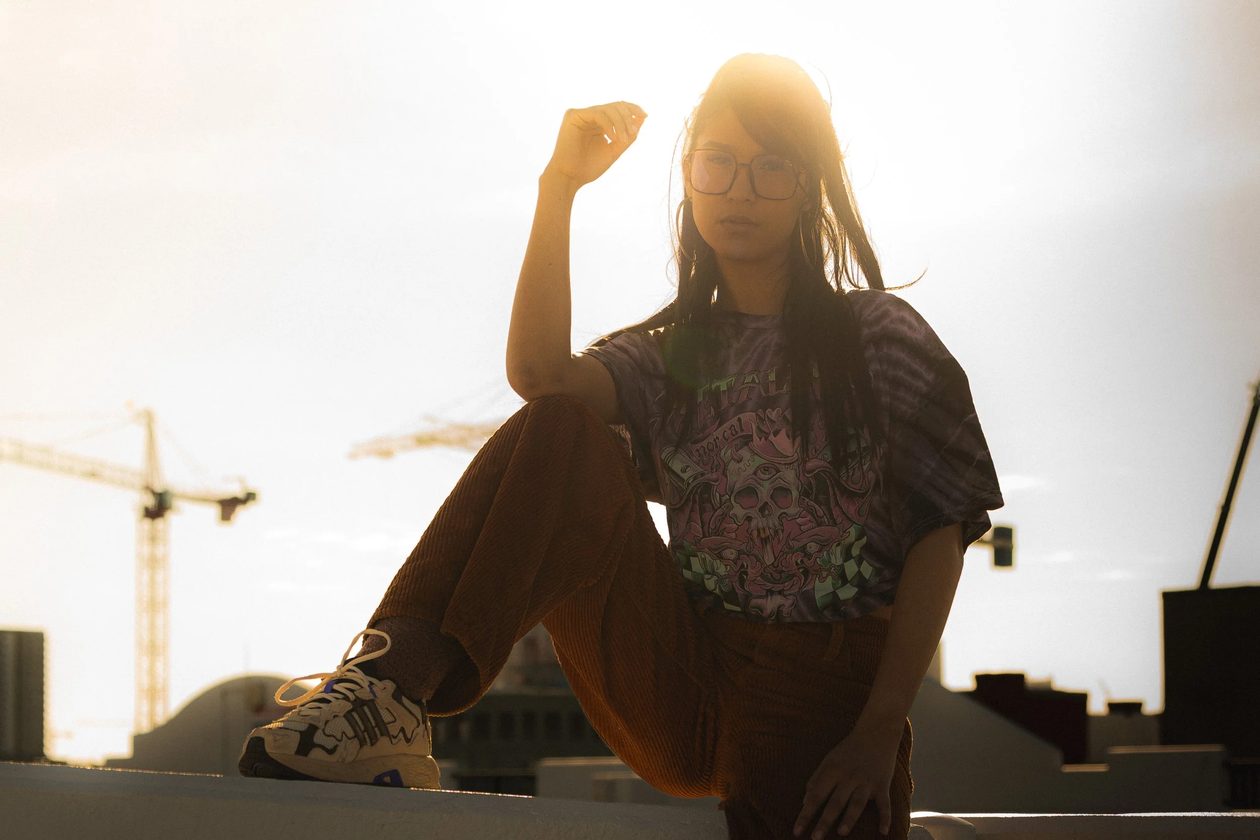 A woman with glasses and long hair posing outdoors against a bright sky with construction cranes in the background.