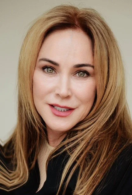 Close-up portrait of a woman with long, reddish-blonde hair, green eyes, and a subtle smile, wearing a dark jacket.