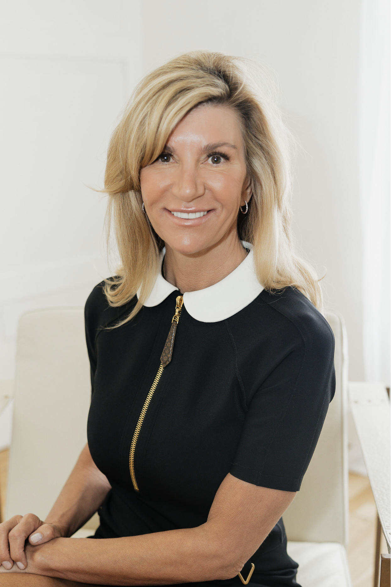A woman with blonde hair wearing a black dress with a white collar and a gold zipper, smiling at the camera in a bright room.
