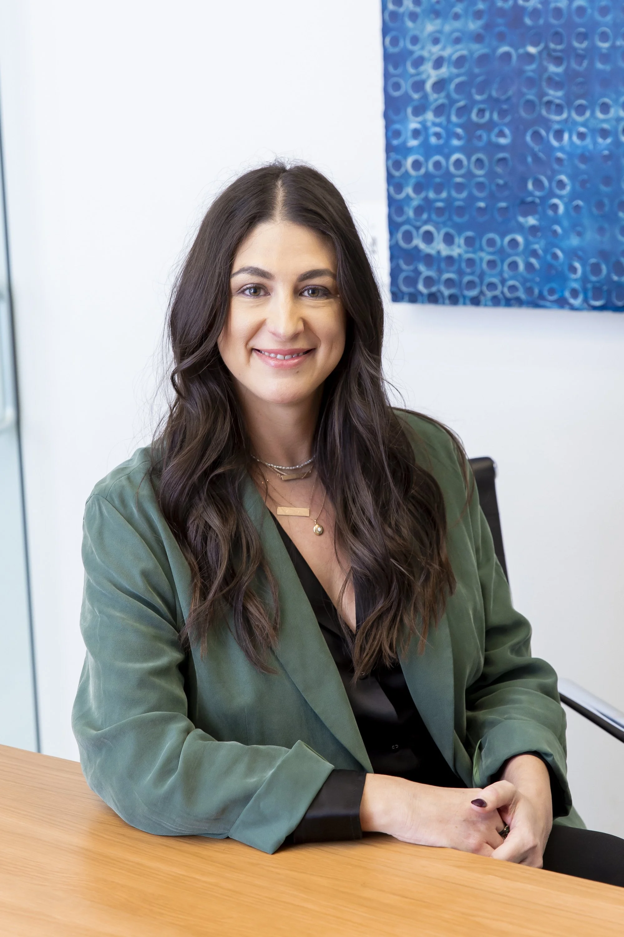 A woman with long dark hair, wearing a green blazer and layered necklaces, sitting at a wooden table and smiling at the camera in an office setting.