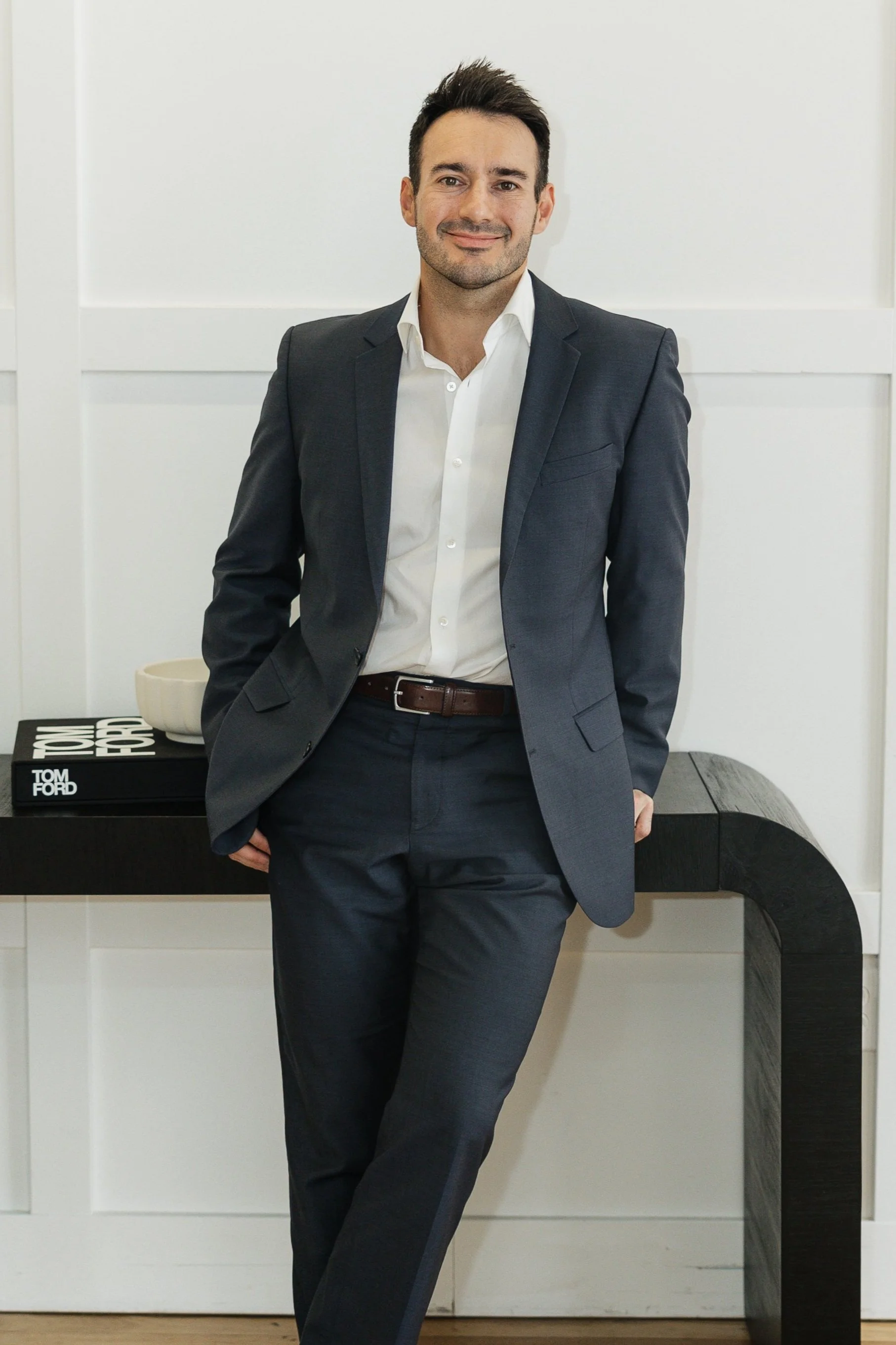 A man in a dark suit and white shirt standing indoors with one hand in his pocket, leaning against a black side table with a stack of books and a beige bowl. The background features white paneled walls.