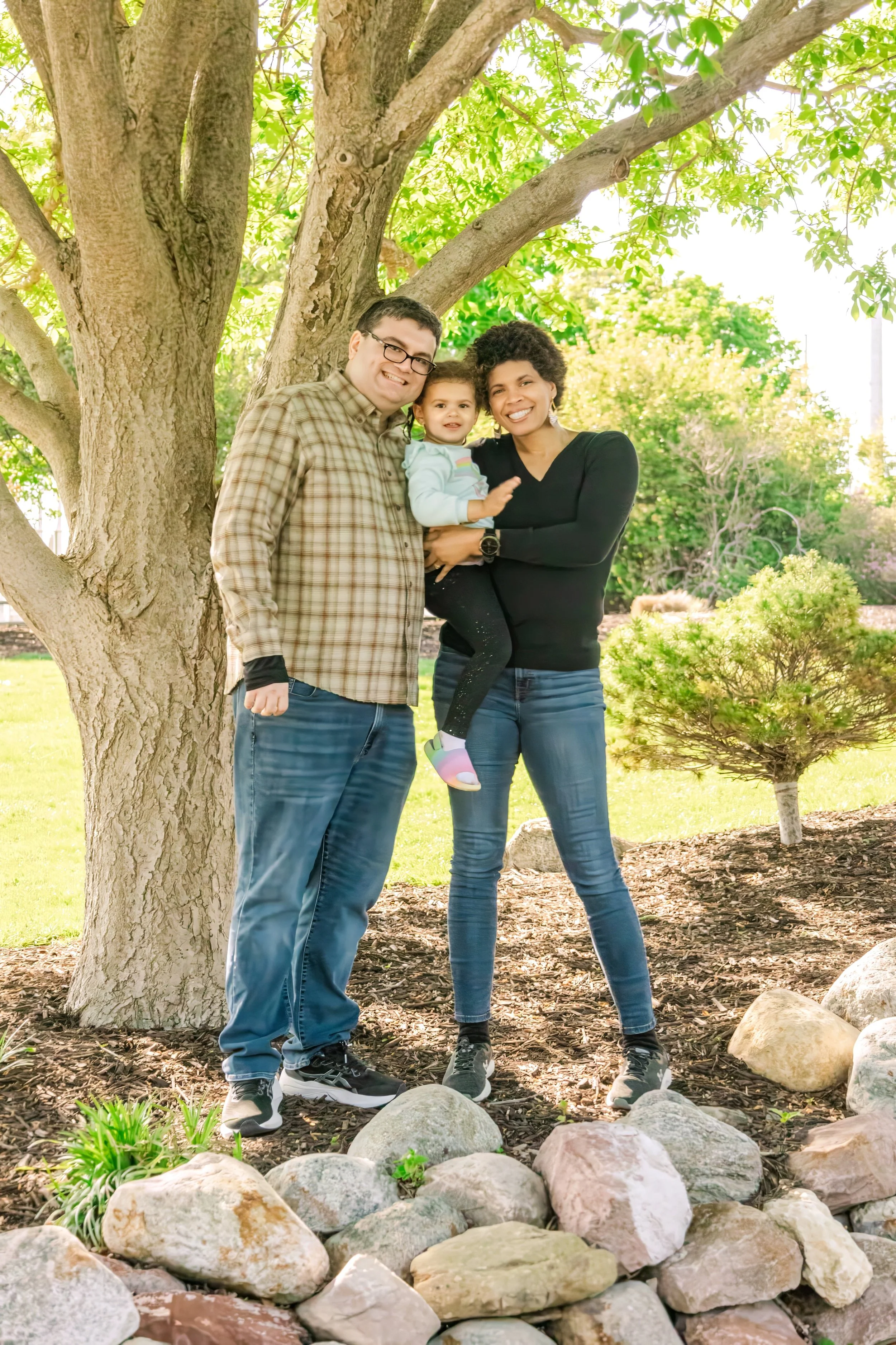 A family of three standing outdoors near a tree, smiling at the camera. The man is wearing glasses and a plaid shirt, the woman is holding a young girl dressed in a light blue jacket and colorful shoes. The background features green trees and sunlight.