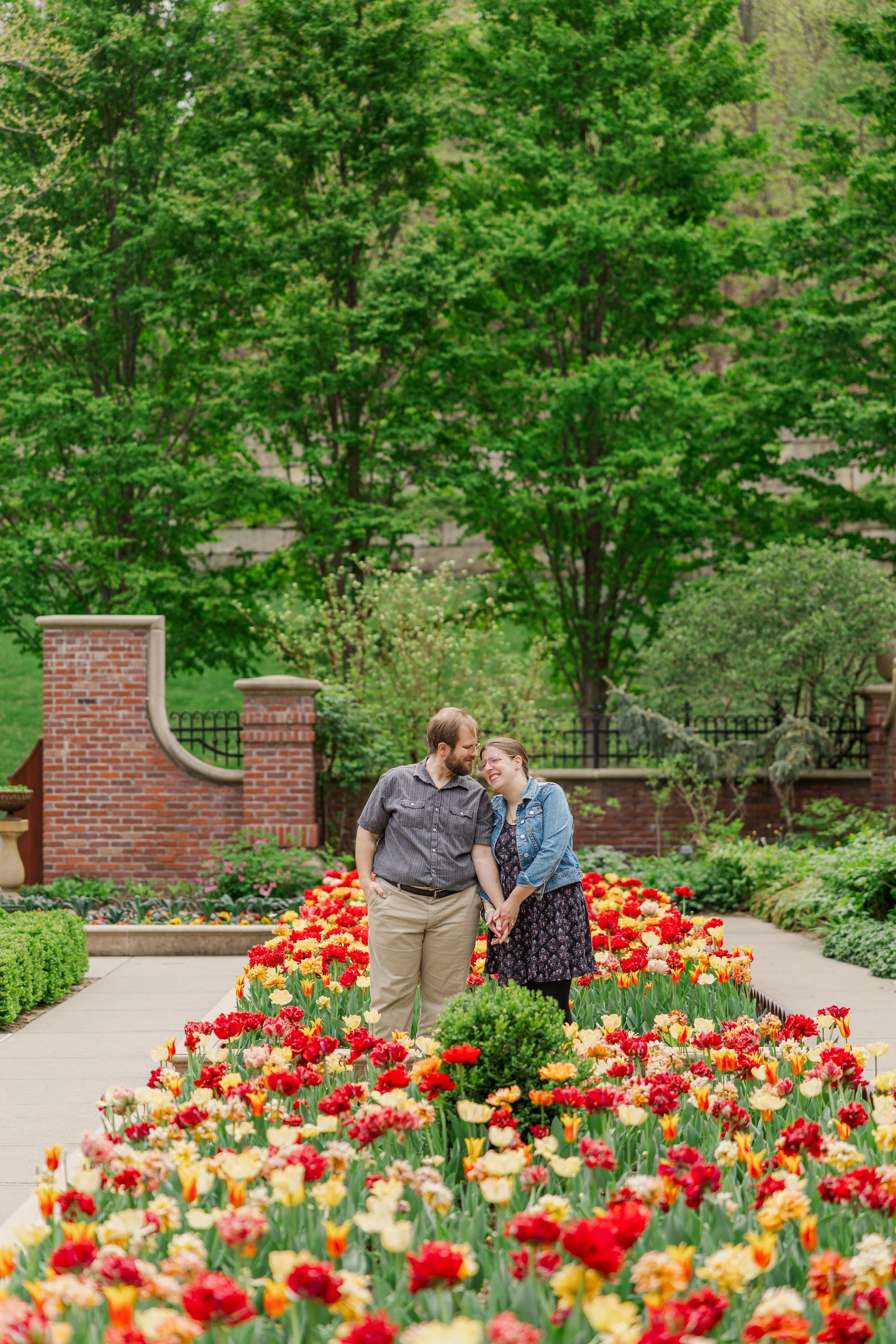 A smiling couple holding hands and leaning their heads together in a garden with colorful tulips and green trees in the background.