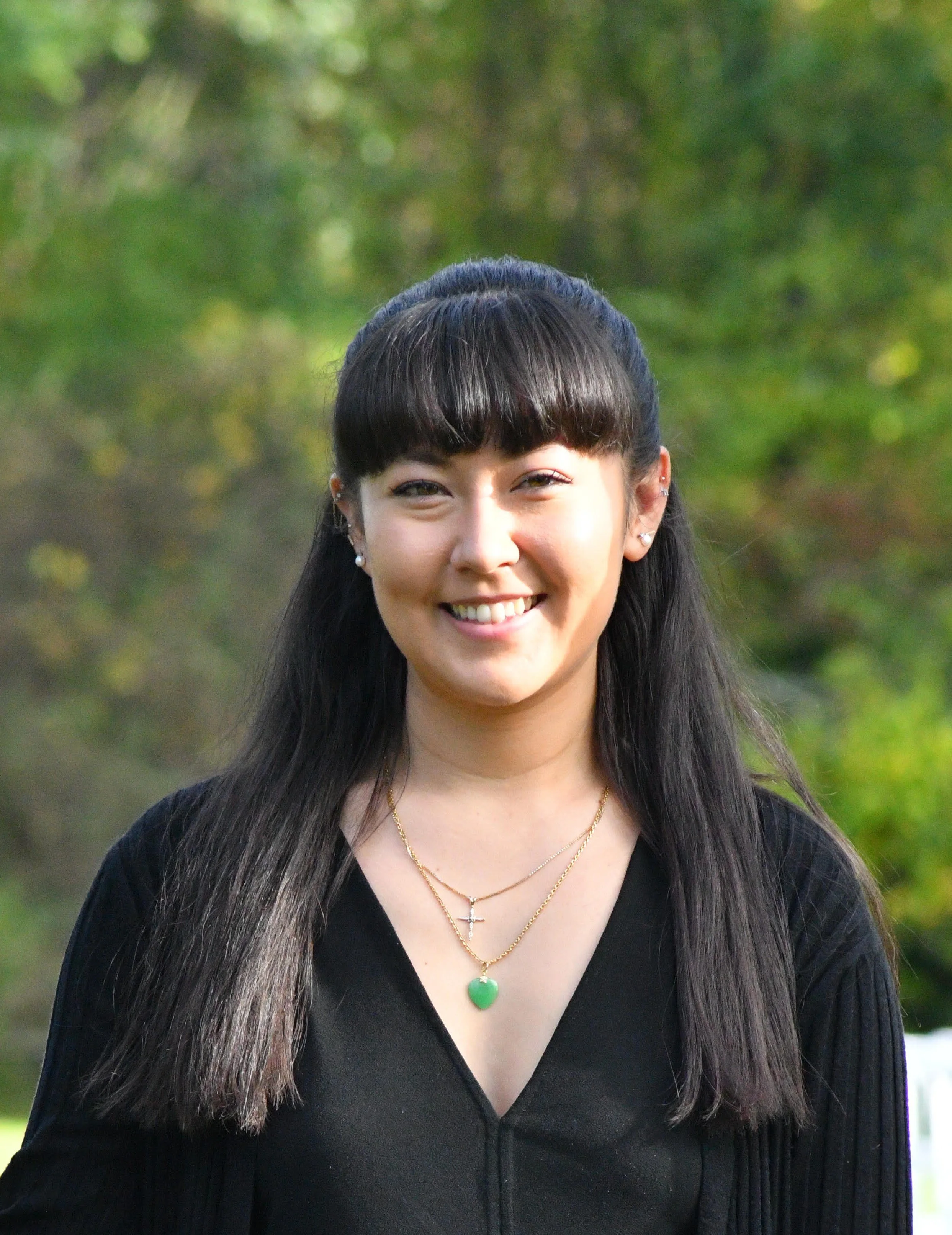 A young woman with long dark hair and bangs smiling outdoors, wearing a black top and layered necklaces, with a green leafy background.