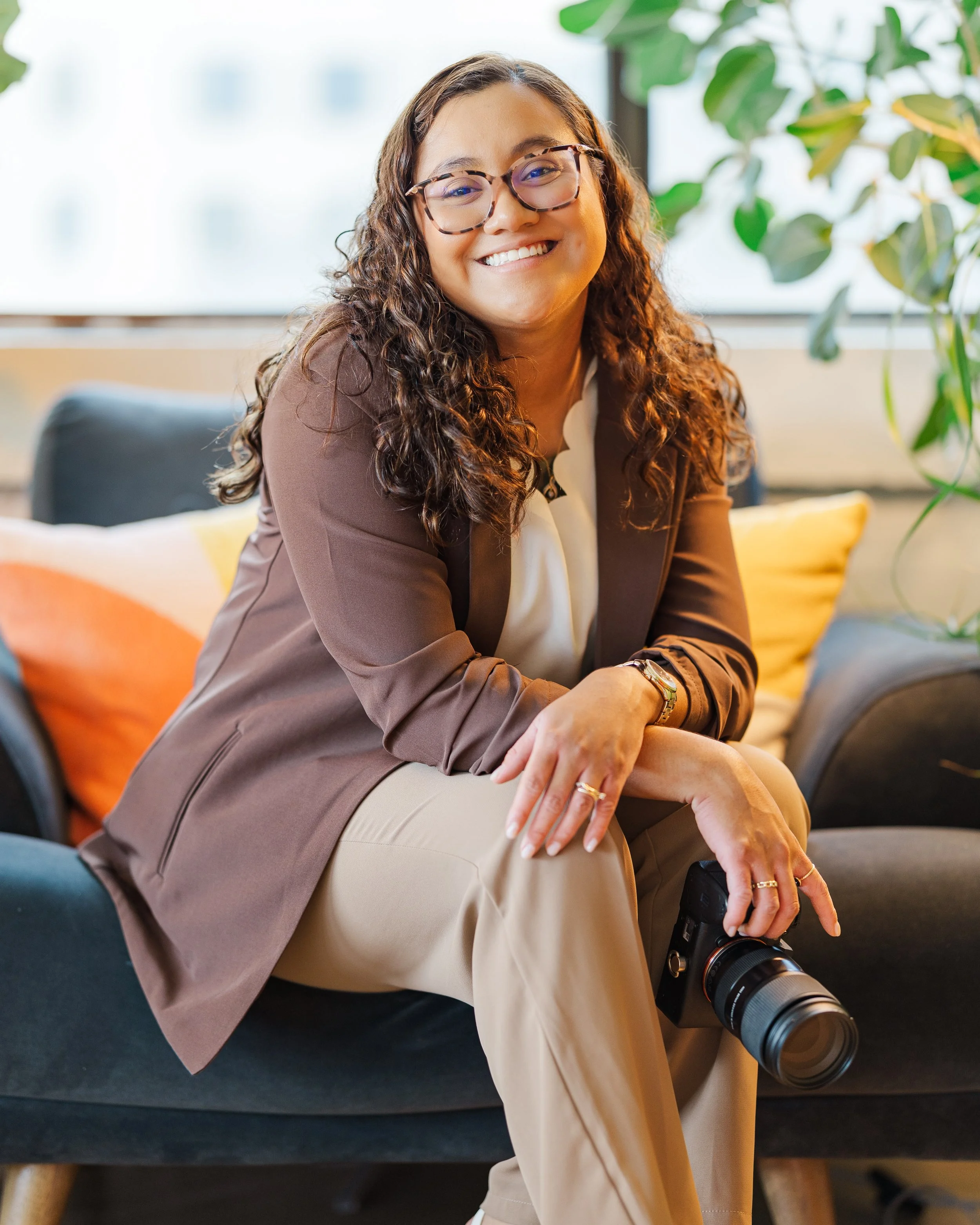 A woman with curly brown hair, glasses, and a warm smile sitting on a dark couch. She is wearing a brown blazer, a white blouse, and beige pants while holding a camera. There are green plants and a window in the background.