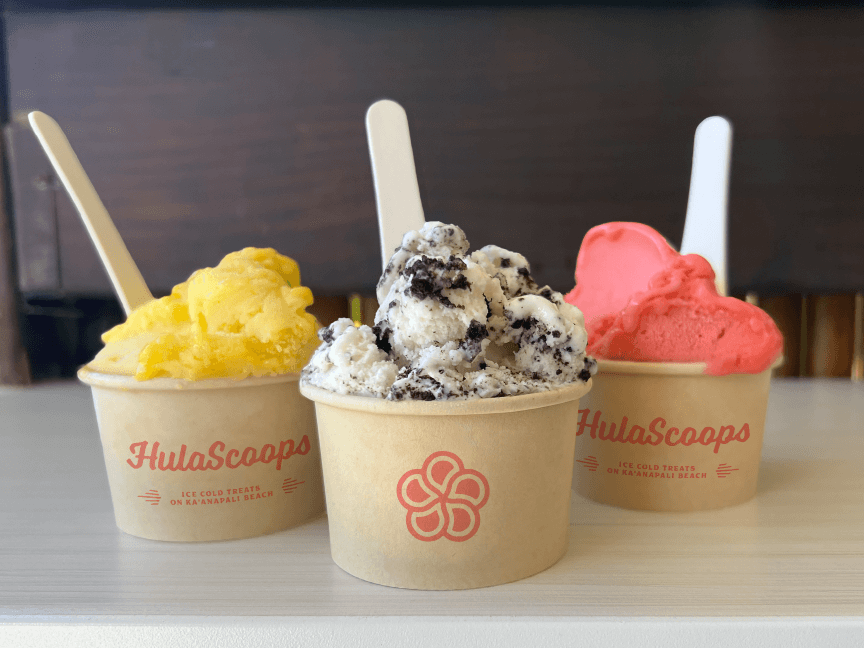 Three paper cups with single size scoops of gelato and sorbetto ice creams sit on a counter at HulaScoops in Maui.