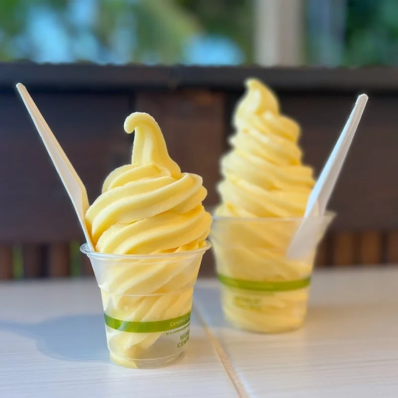 Two cups filled with bright yellow pineapple Dole Whip sit on a tile countertop at HulaScoops in Maui with palm trees in the background.