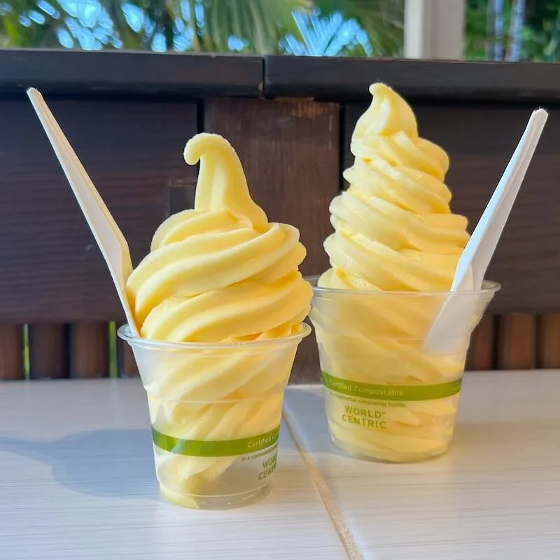 Large and small clear cups filled with yellow swirls of pineapple Dole Whip sit on the tile counter at HulaScoops in Maui.