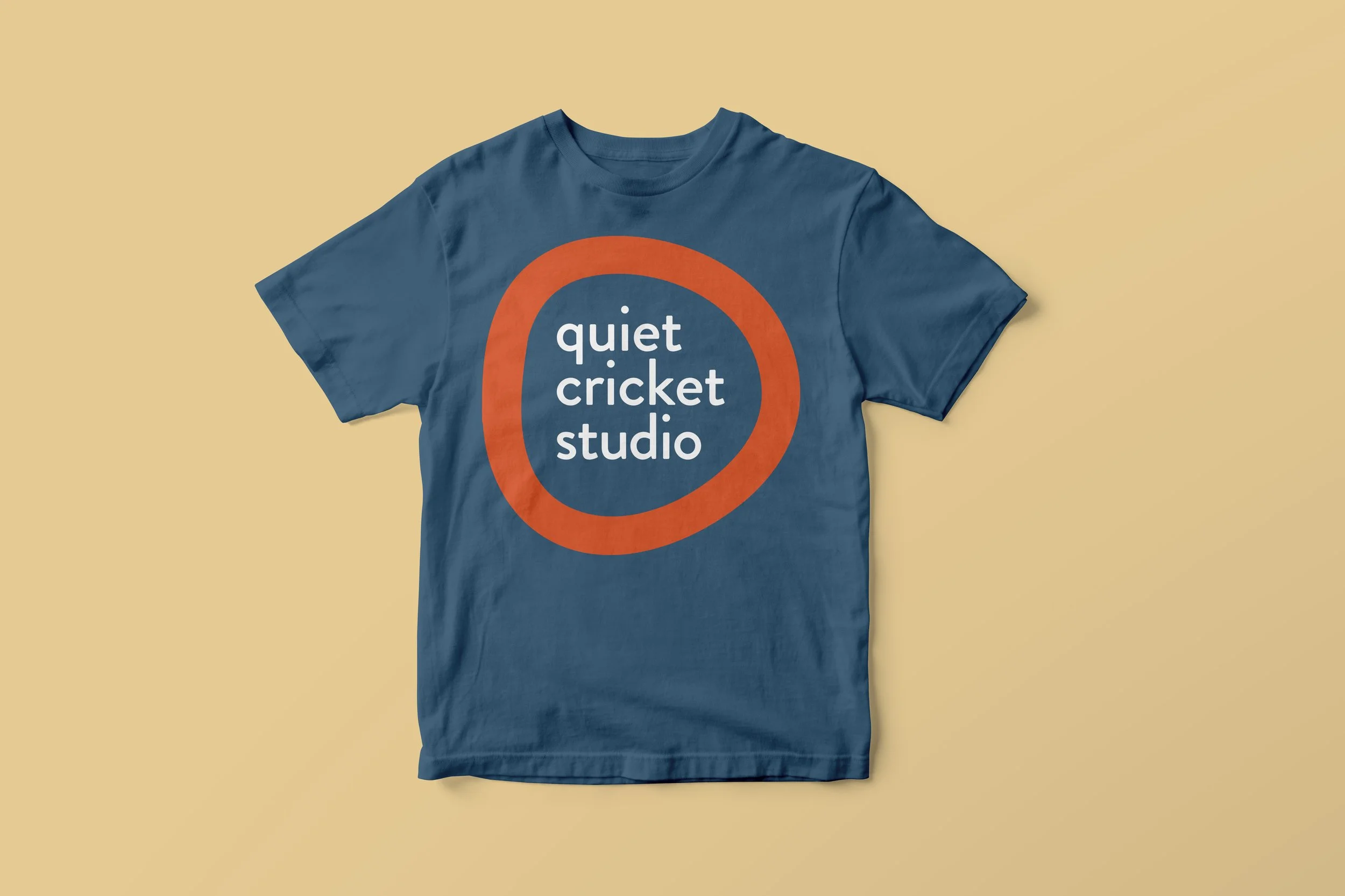 Blue T-shirt with orange circle and white text reading 'quiet cricket studio' on a light orange background.