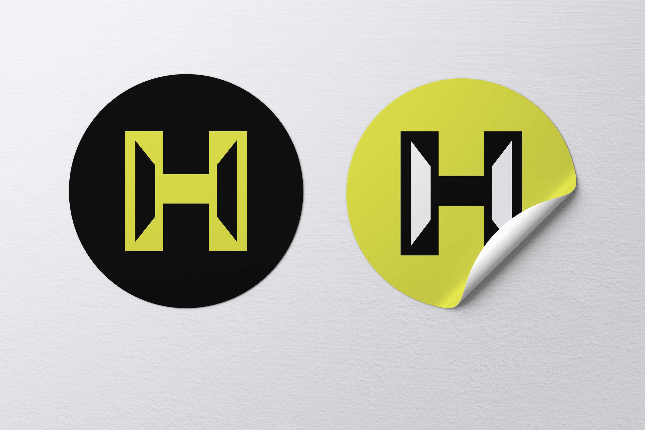 Two round stickers with a stylized 'H' logo, one black with yellow-green logo and the other yellow-green with a black and white logo, stuck on a wall, one sticker slightly peeling off.