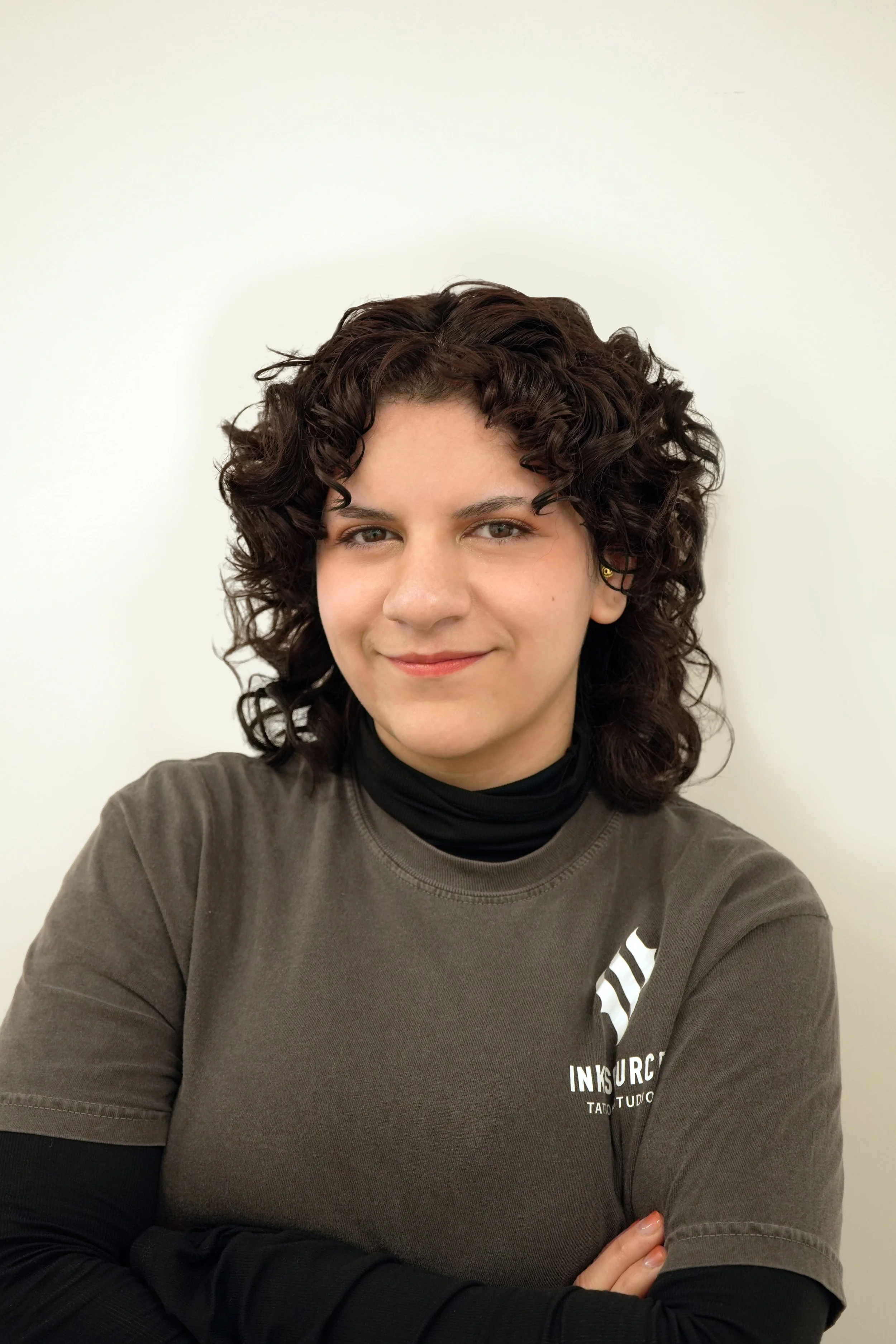 A woman with curly dark hair smiling, wearing a dark gray sweatshirt and a black long sleeve shirt underneath, standing against a plain white wall.