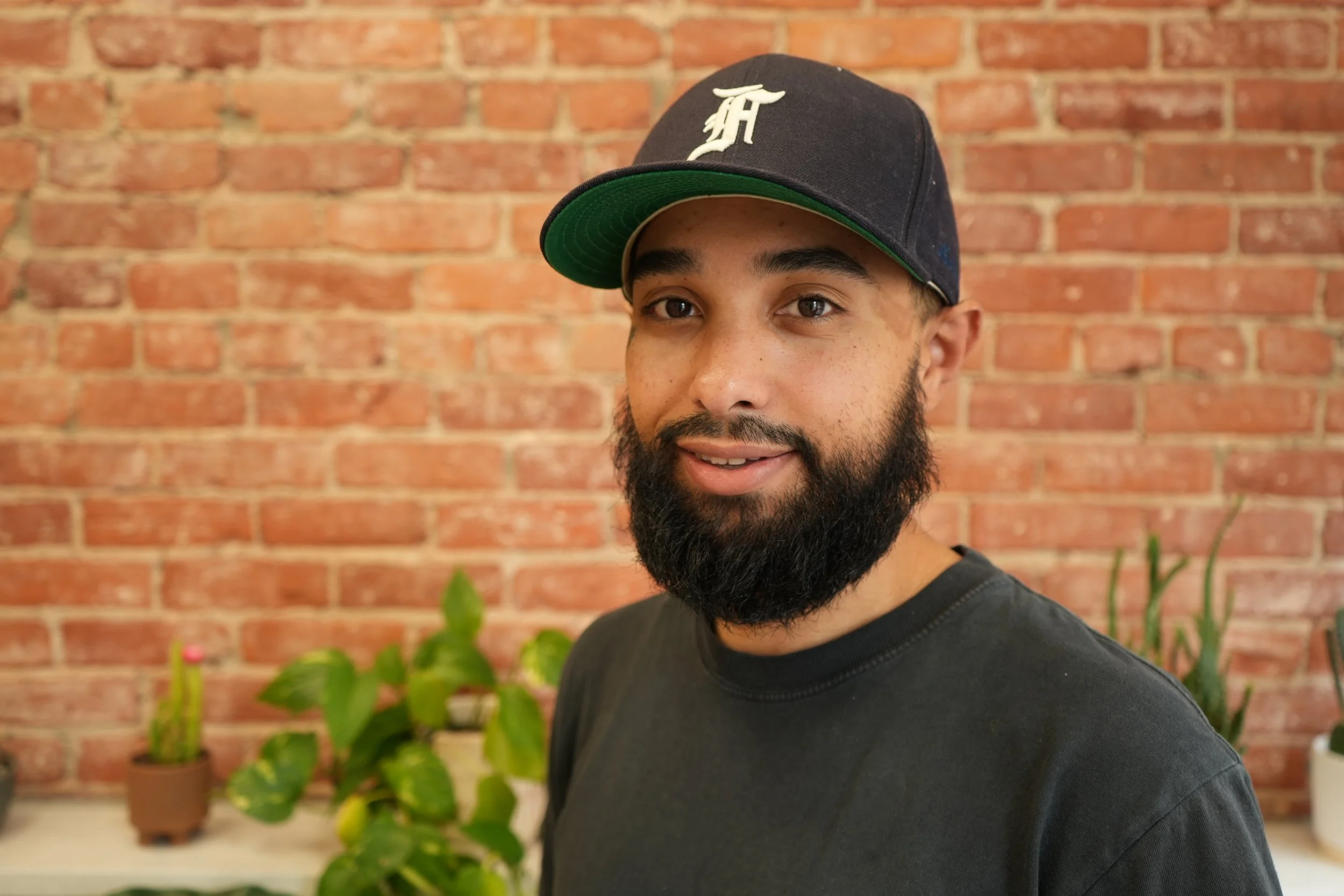 A man with a beard wearing a black baseball cap with a white 'D' logo and a black T-shirt, smiling in front of a red brick wall and green potted plants.