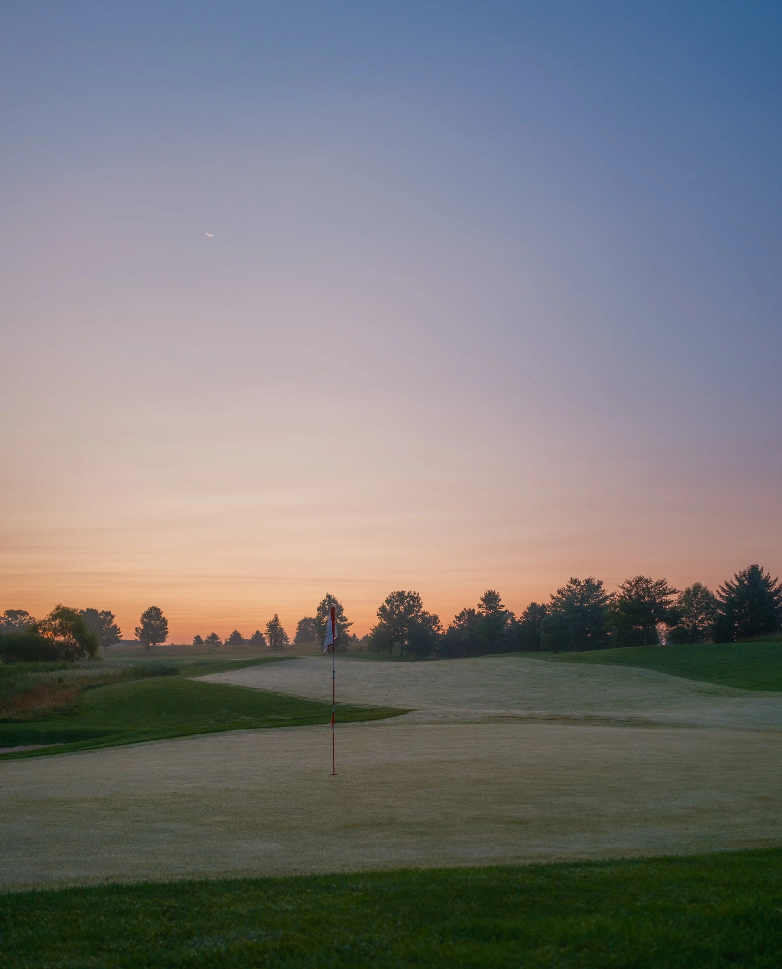 A golf course at sunset with a flag on a green and trees in the background.