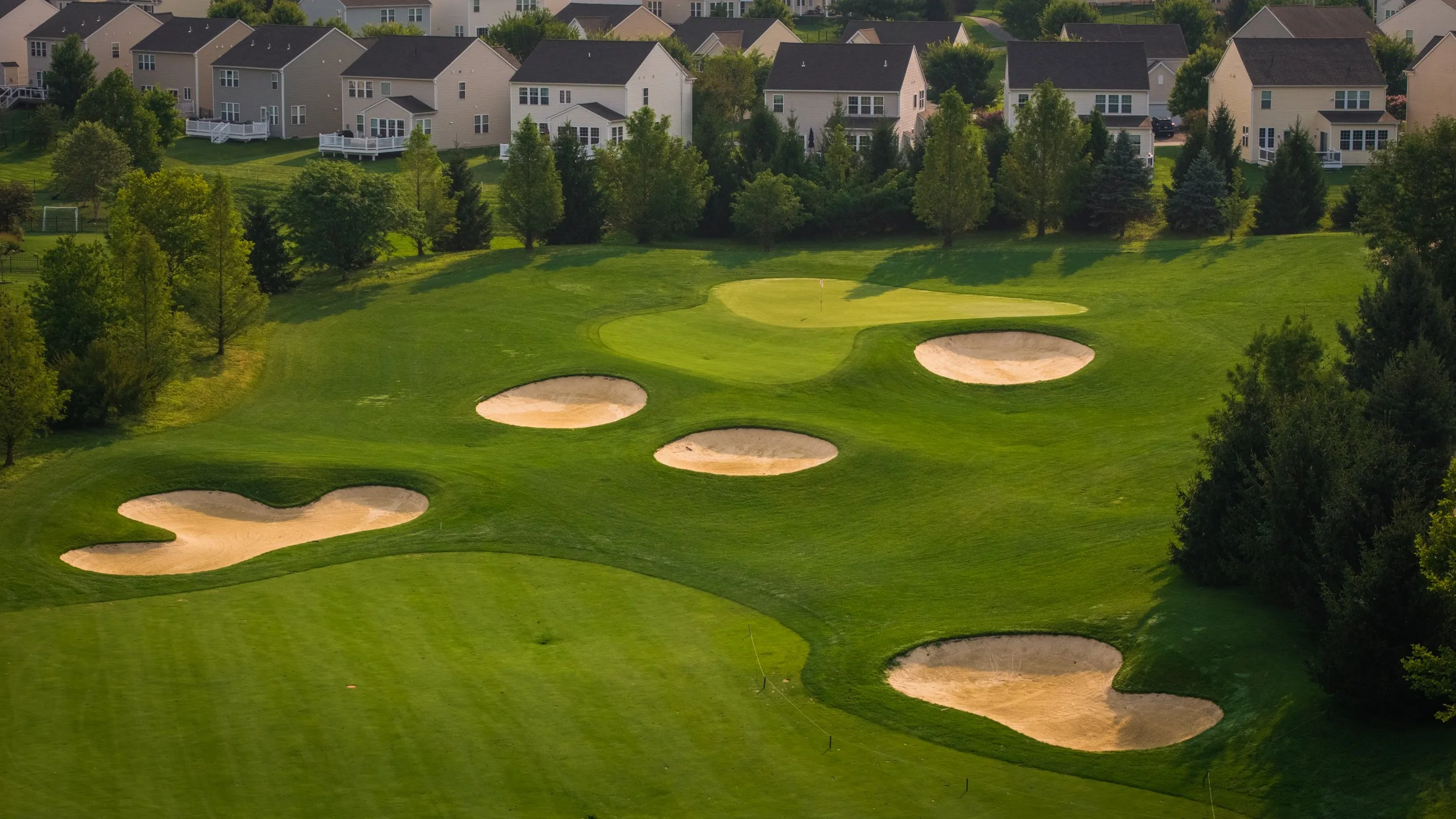 A golf course with several sand traps and a green, surrounded by trees and residential houses in the background.