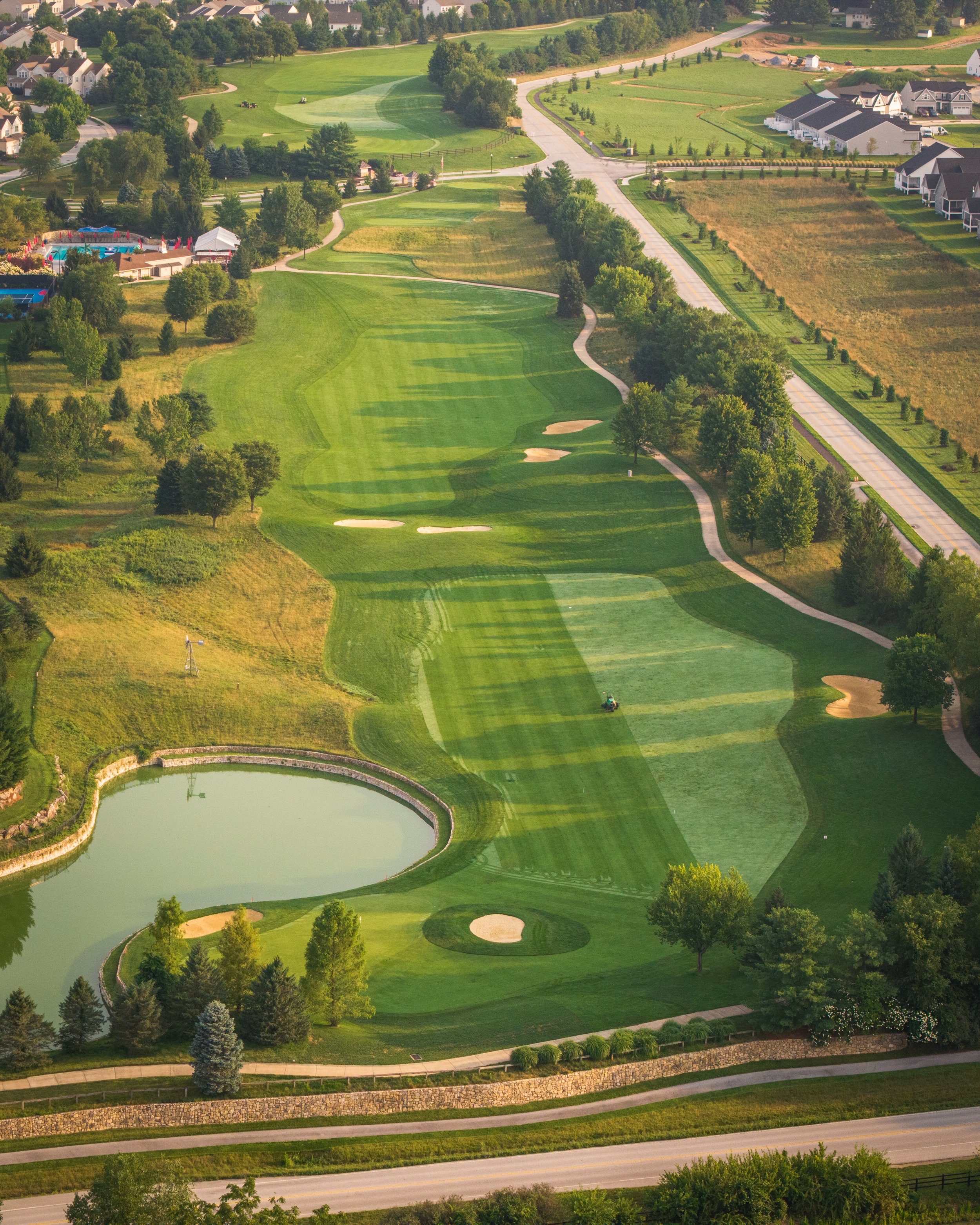 Aerial view of a golf course with well-maintained greens, bunkers, sand traps, and a pond. Surrounding the course are trees, pathways, and residential houses in the background.