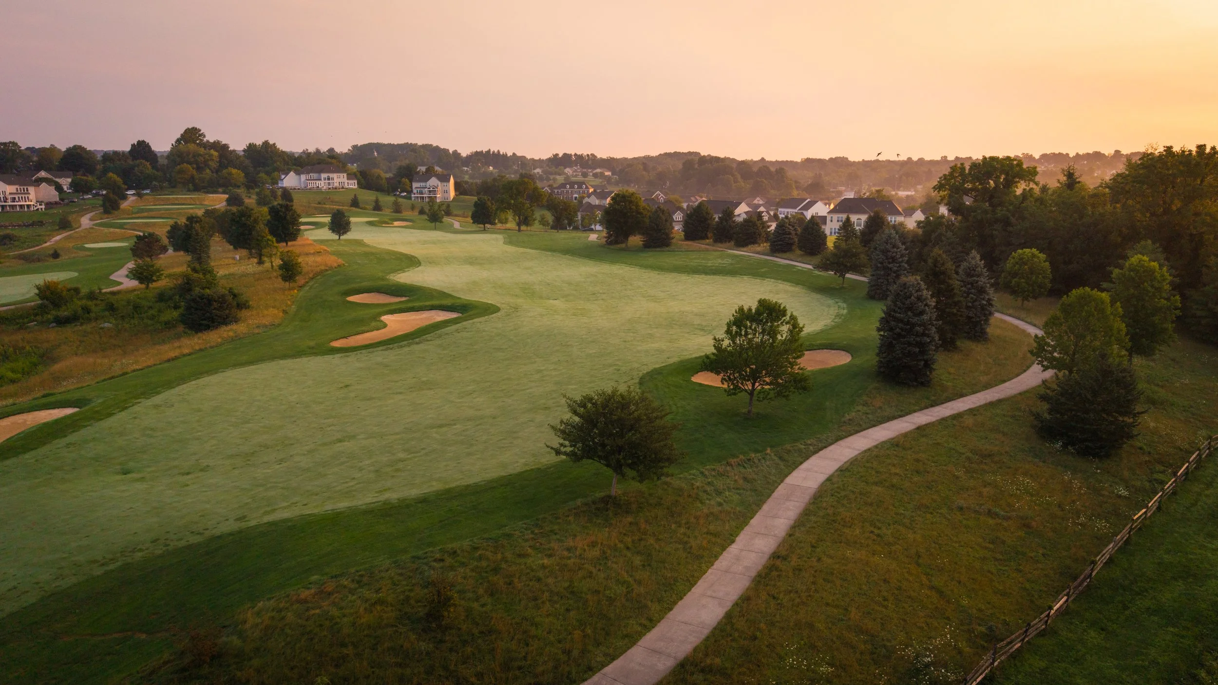 Sunset view of a golf course with green fairways, sand traps, trees, and houses in the background.