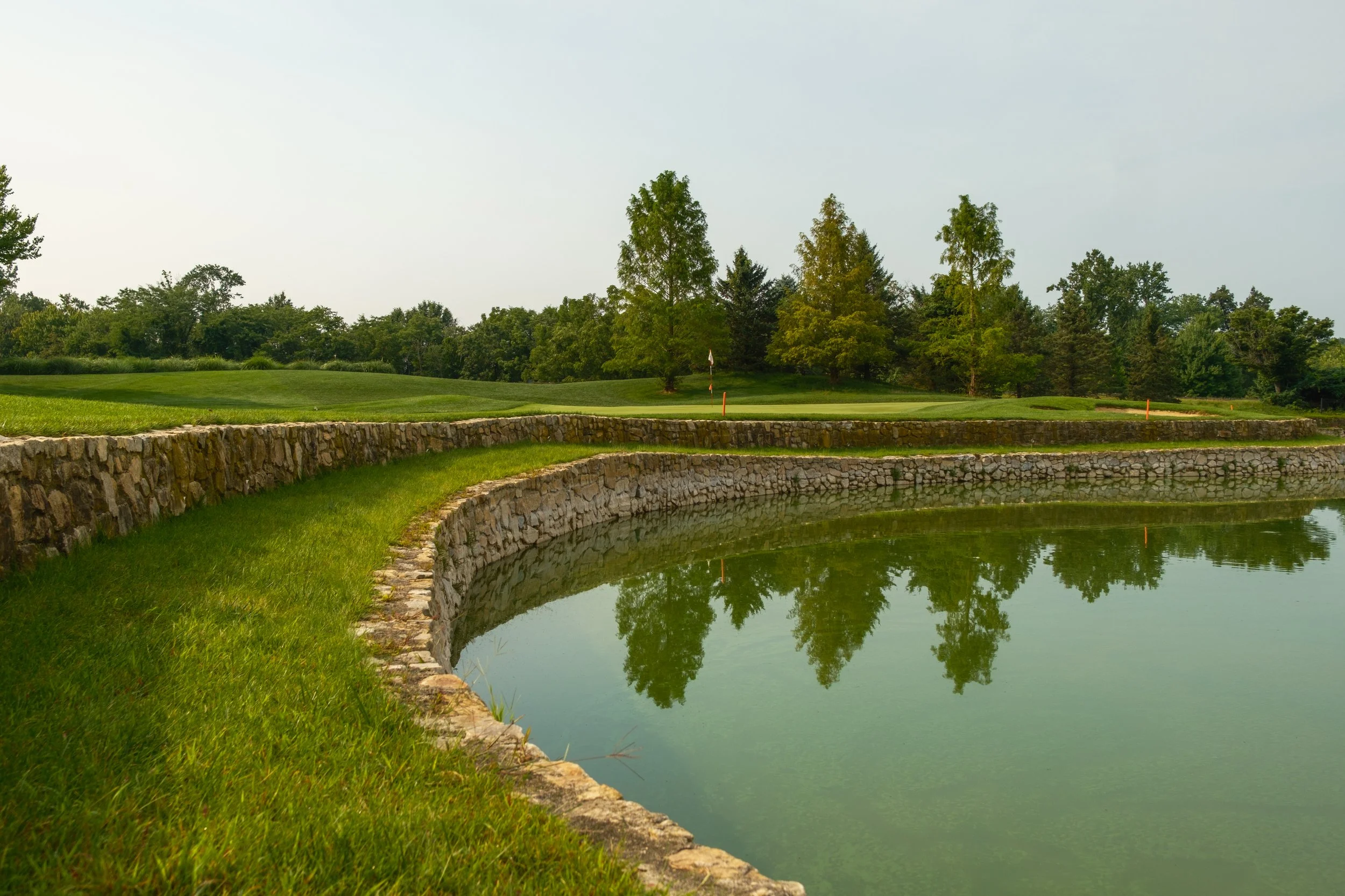 A golf course with a small pond, lush green grass, trees in the background, and a cloudy sky.
