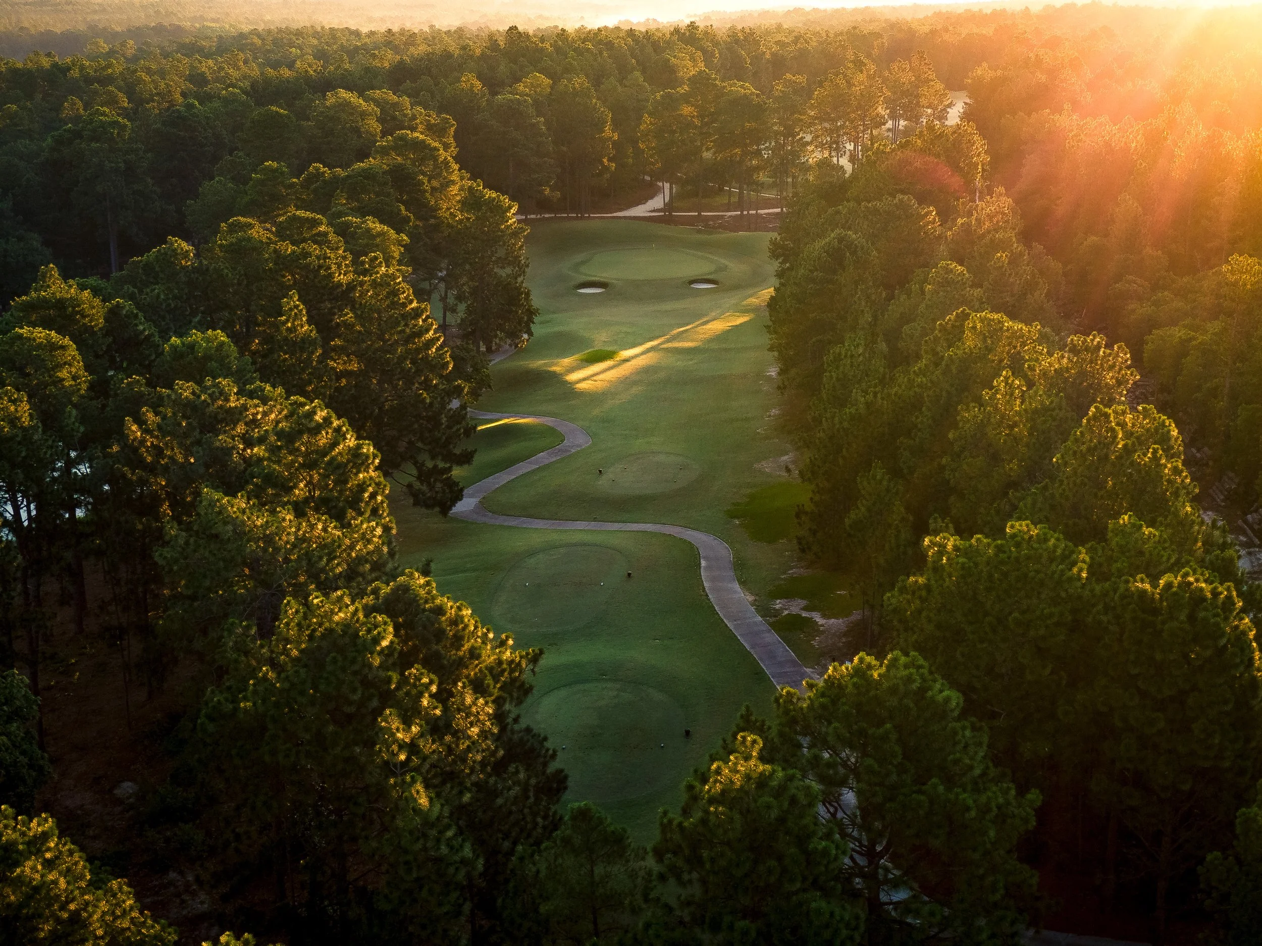 Aerial view of a golf course surrounded by trees with the sun setting in the background.