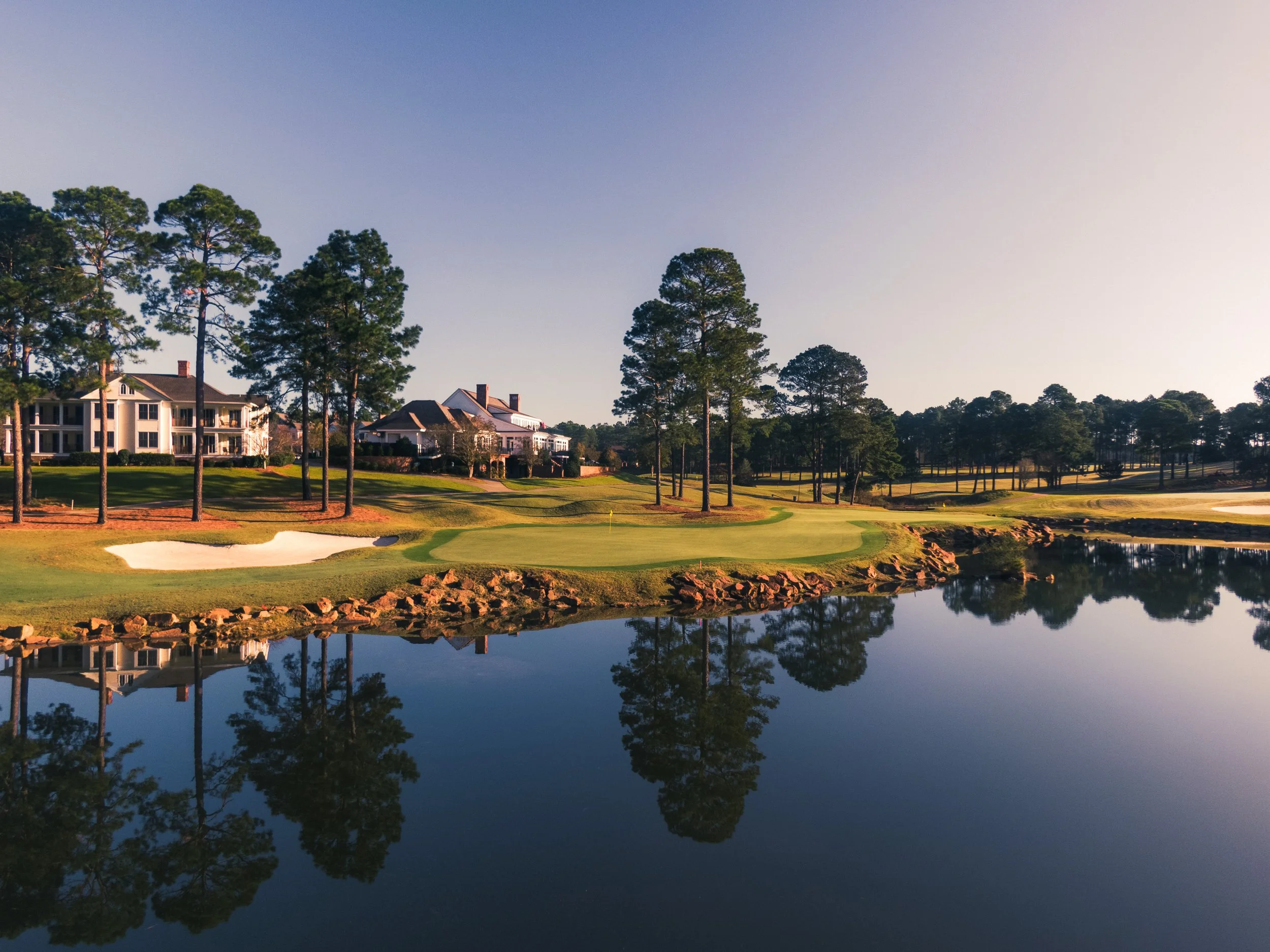 A golf course with a water hazard, trees, and residential houses in the background during sunset, with reflection of the scene in the water.