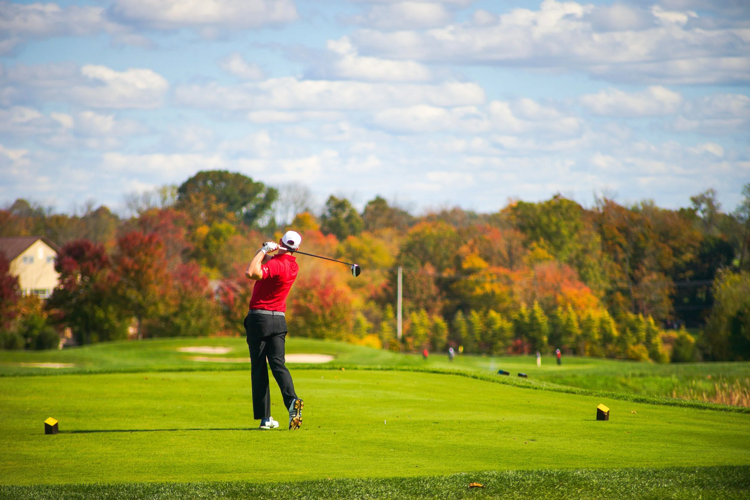 A golfer in a red shirt and black pants is swinging a golf club on a golf course, with trees displaying autumn colors and a partly cloudy sky in the background.
