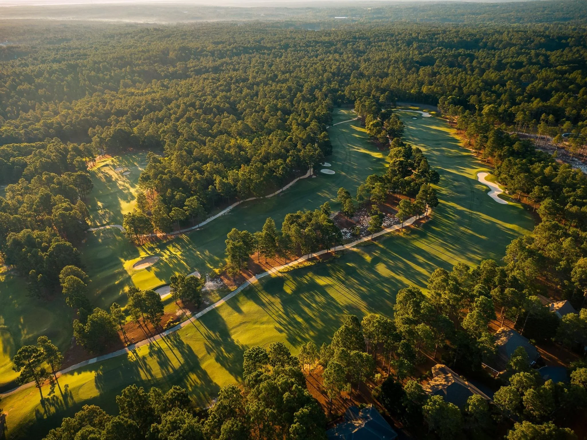 An aerial view of a lush green golf course surrounded by dense forest and trees, with several sand traps and curved fairways visible.