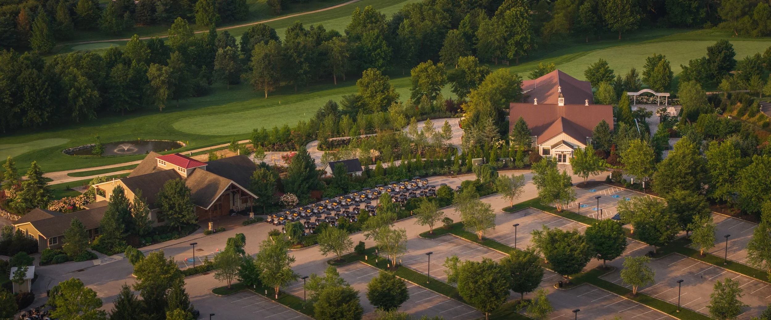 Aerial view of a golf course and parking lot surrounded by trees, with buildings and a pond nearby.