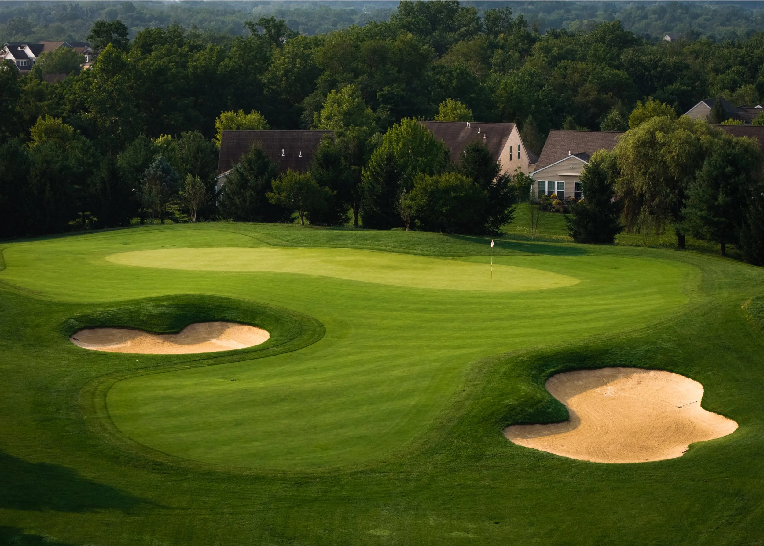 A golf course with multiple sand bunkers and a putting green, surrounded by trees and residential houses in the background.