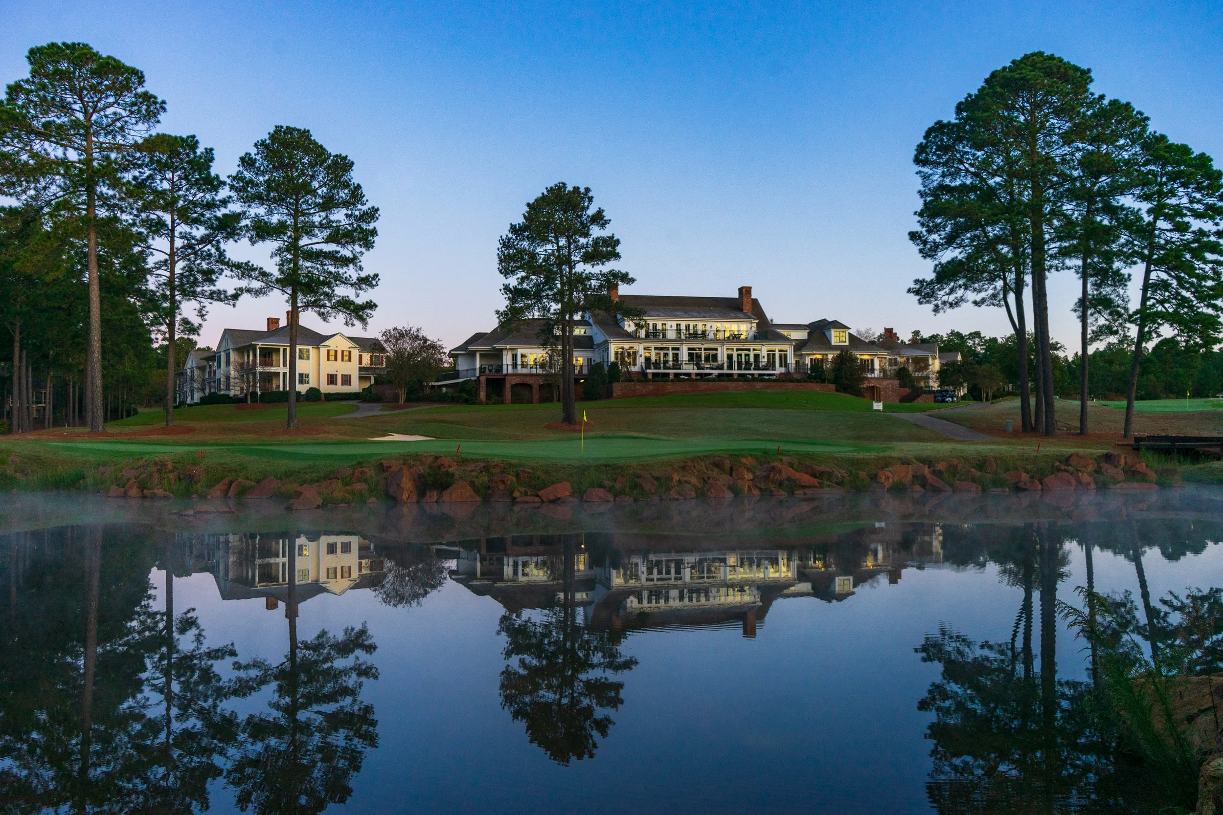 A large house with a porch and multiple stories on a hill, surrounded by trees, with a golf course in front and a pond reflecting the house and trees at dusk.
