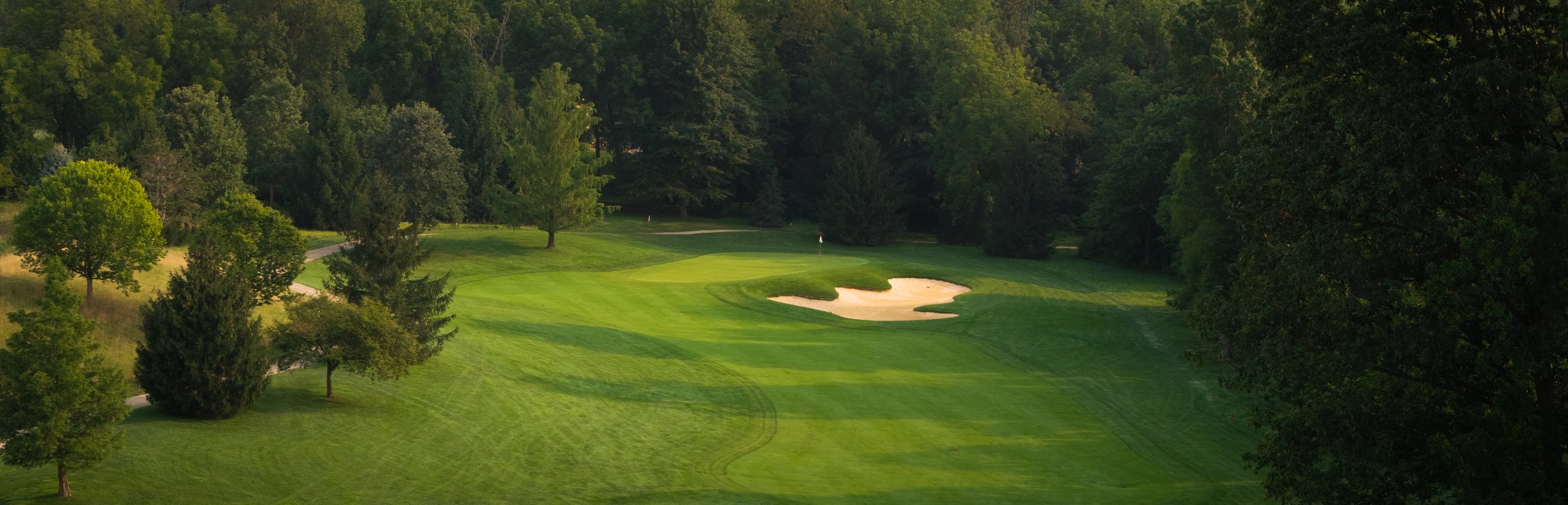 View of a golf course fairway with a sand trap, surrounded by lush green trees.
