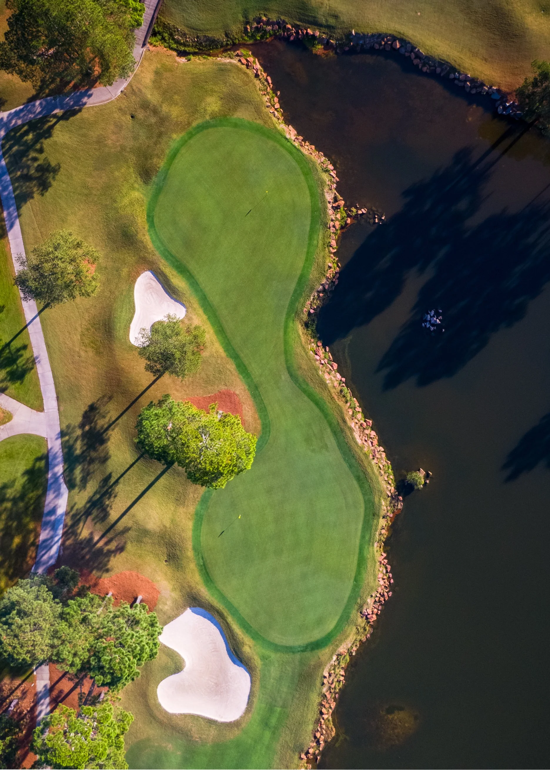 Aerial view of a golf course with two putting greens, sand bunkers, trees, and a large pond along the right edge.