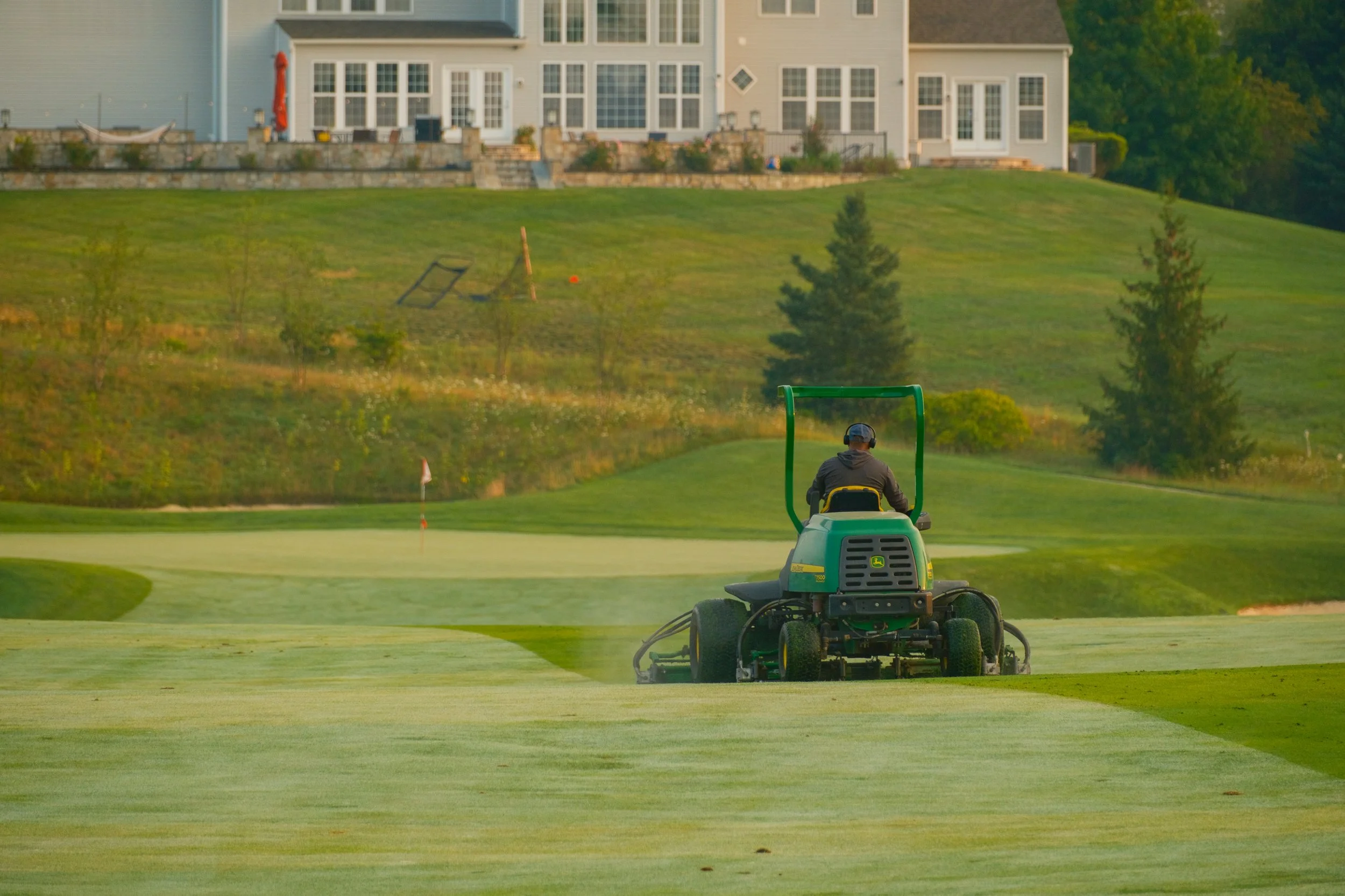 A person riding a green lawn mower on a well-maintained grassy area, with a row of houses and trees in the background.