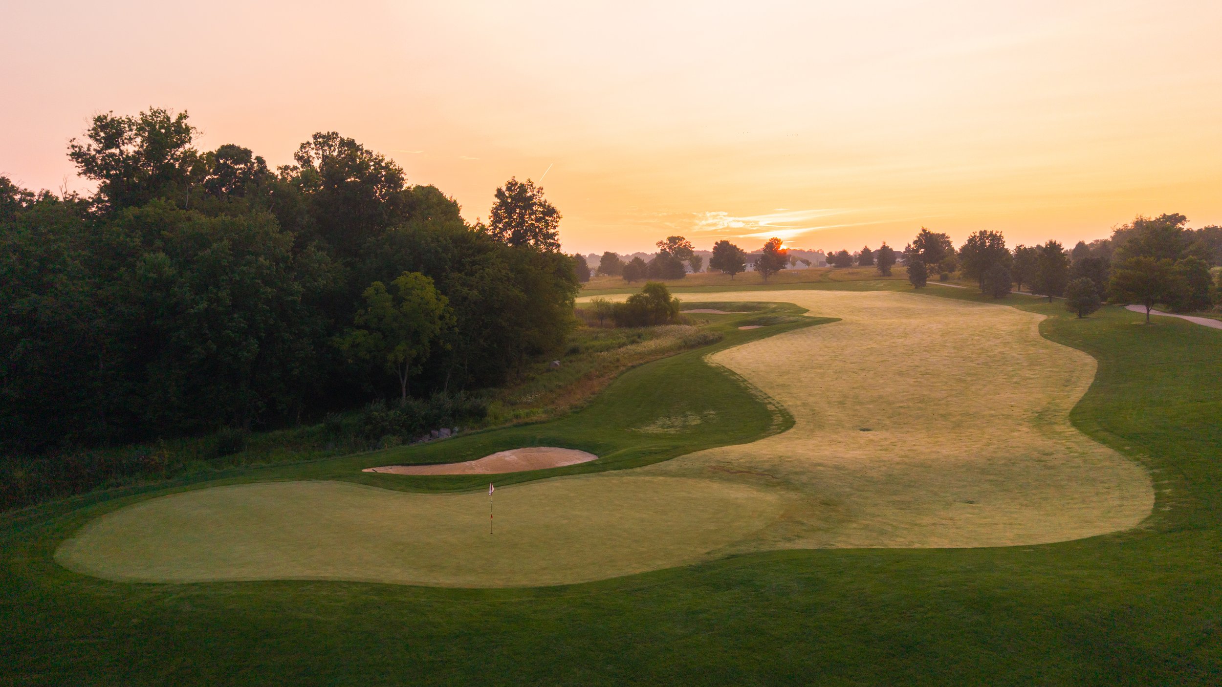 A golf course during sunrise with lush green fairways, sand bunkers, and distant trees under a pastel sky.