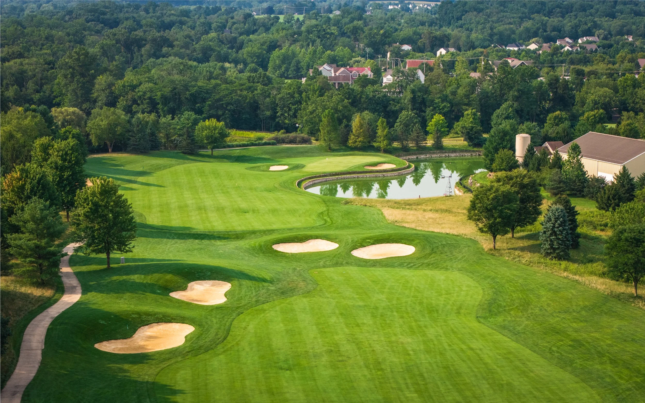 An aerial view of a golf course with neatly maintained green grass, multiple sand bunkers, a water hazard, surrounded by trees and residential houses in the background.
